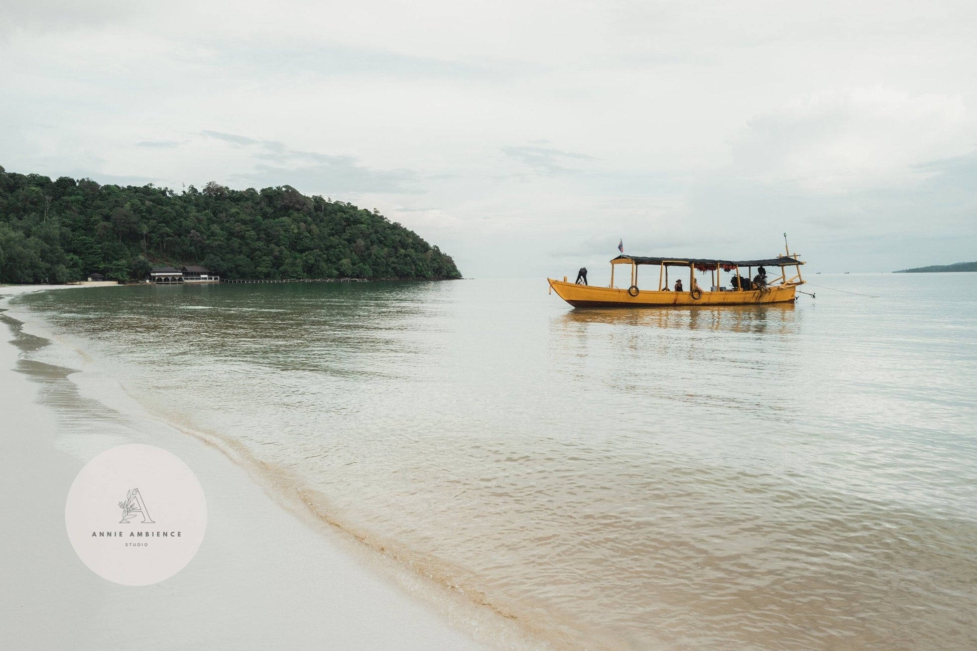 A Koh Rong boat on calm waters near a sandy beach, with a tree-covered hill in the background.
