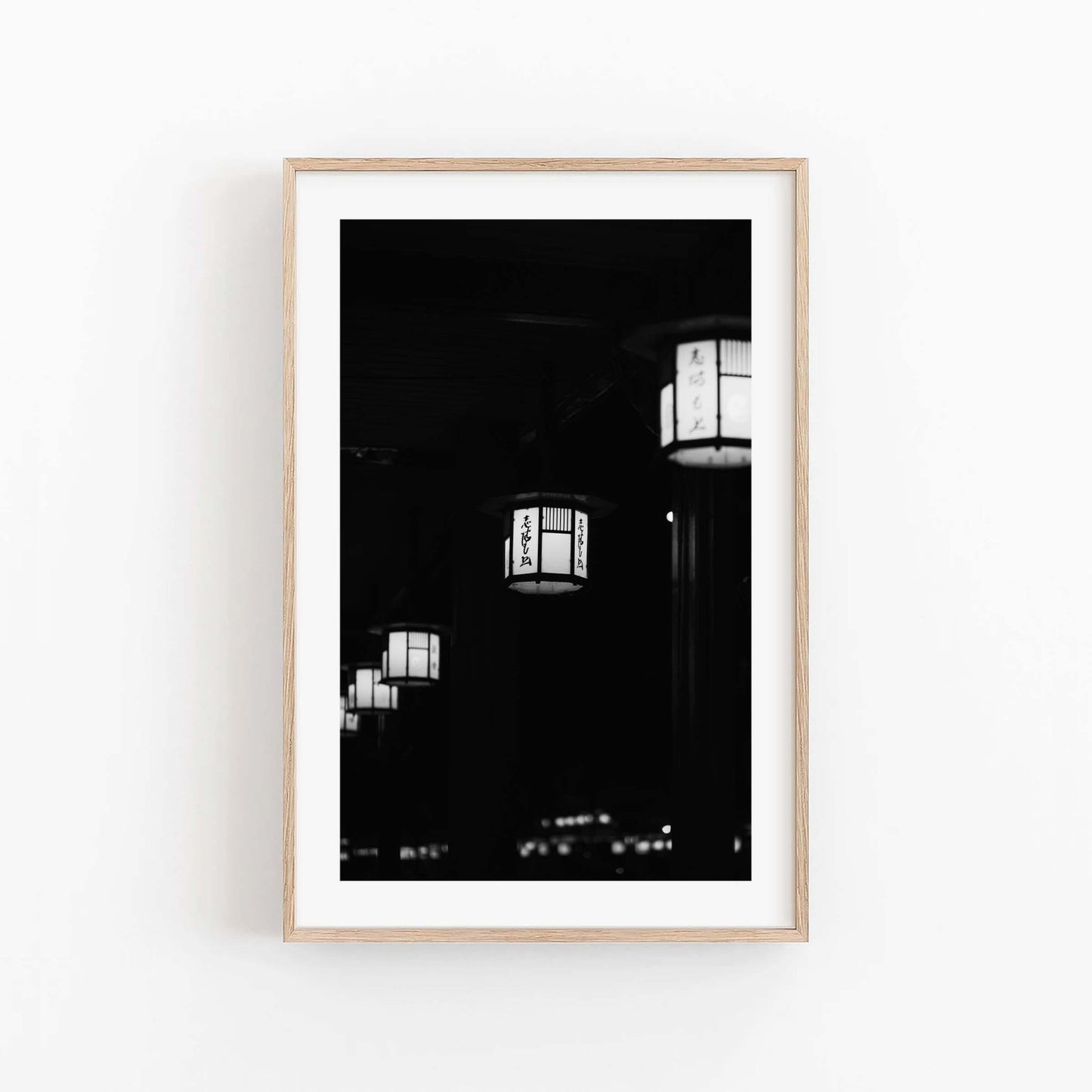 Kyoto Lanterns II Black and White, framed in light wood, features Japanese lanterns on a white wall.