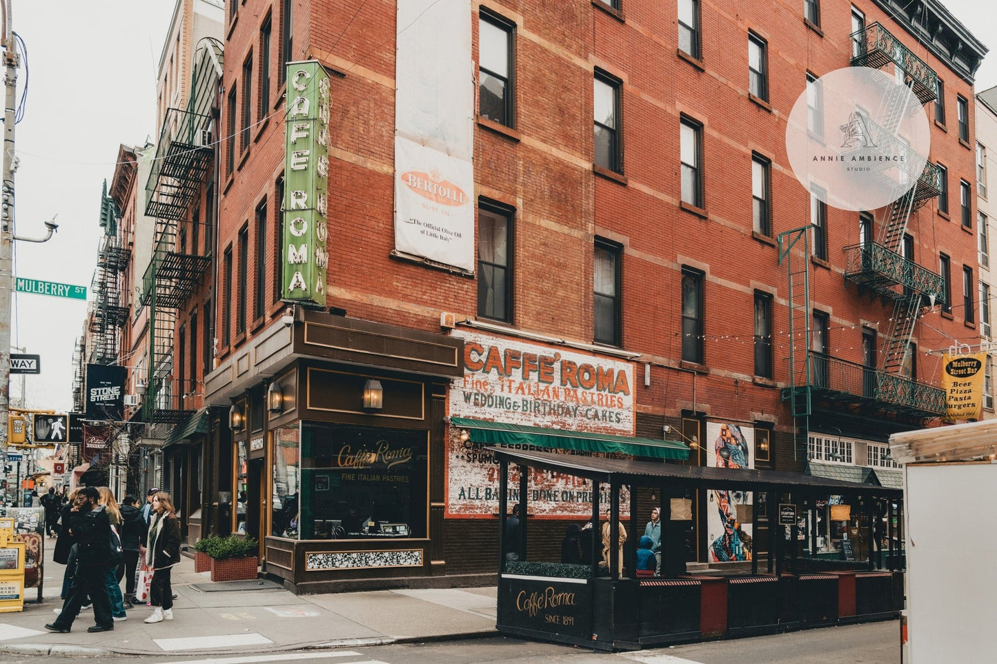 Street view of Little Italy II, a vintage brick building with neon signs and outdoor seating. Pedestrians walking nearby.