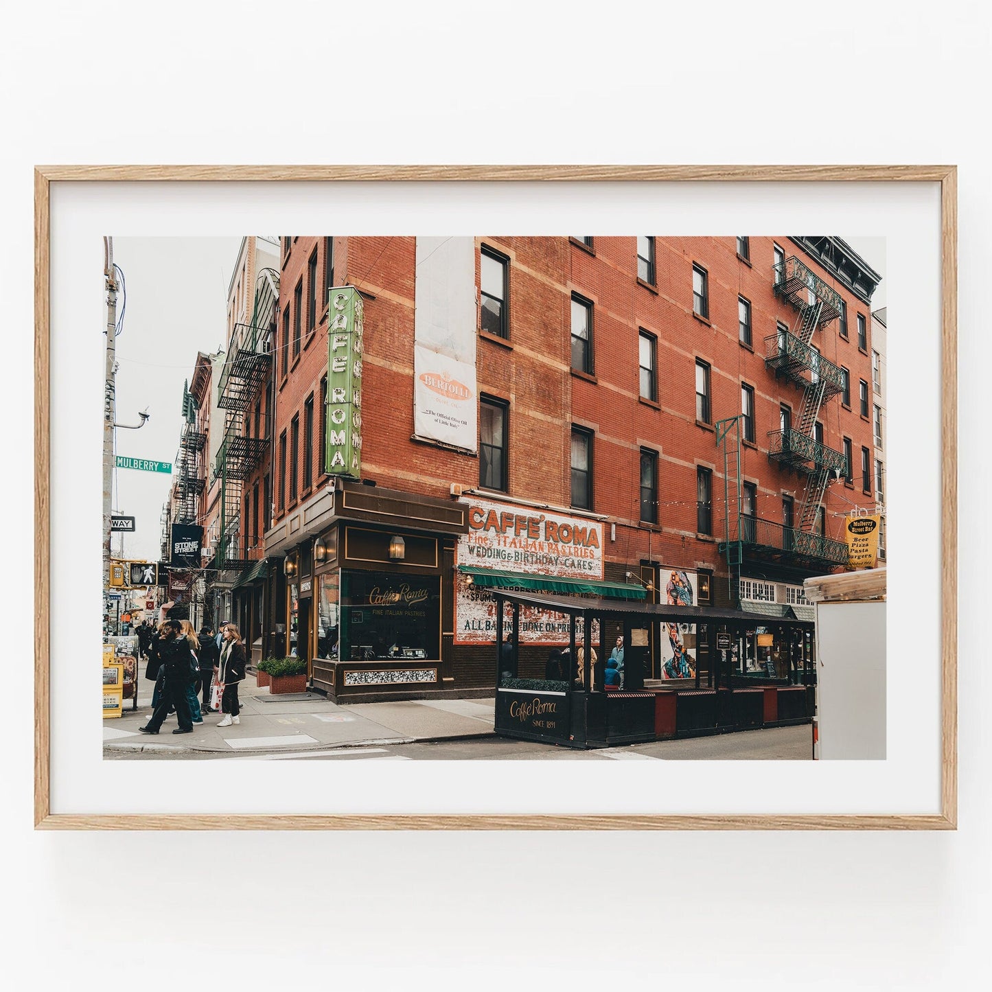 Little Italy II: A framed street scene featuring red brick buildings, a café, and people strolling by a city corner.