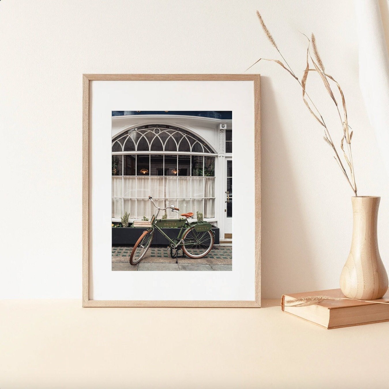 Framed photo London Bike features a vintage bicycle by an arched window, with a vase and books on a beige surface.