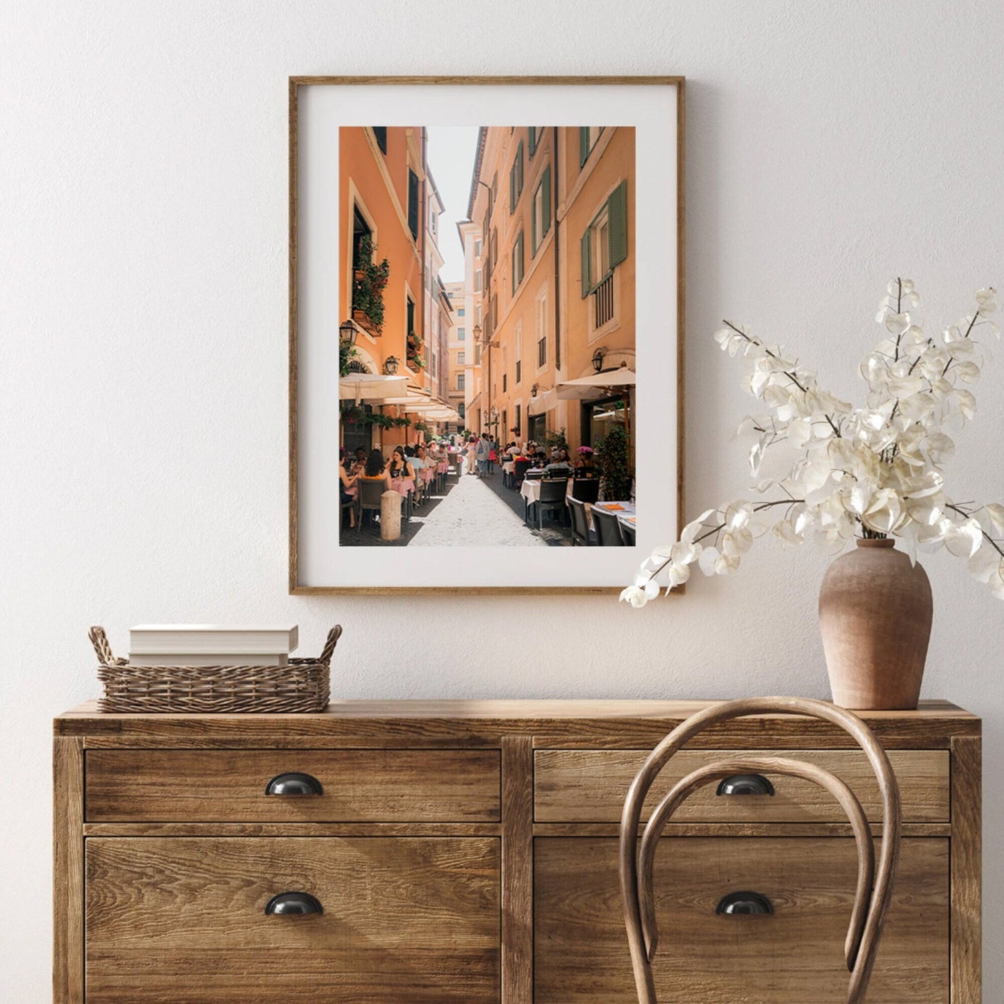 The Lunch in Rome artwork hangs above a wooden dresser with a wicker basket and vase of flowers.