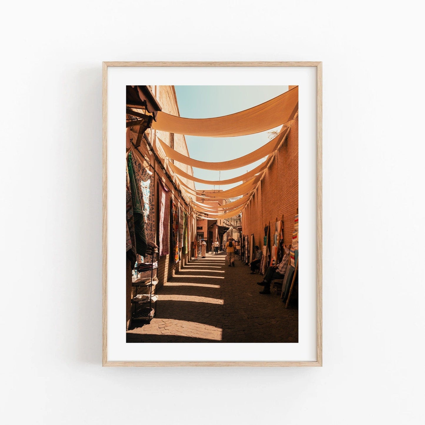Framed Marrakesh Souk I photo: sunlit market alley with hanging fabric shades and colorful textiles.