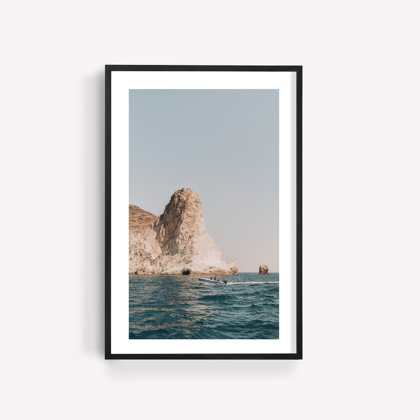 Framed photo Mediterranean Adventure features a rocky seaside cliff, calm blue water, and clear sky on a white wall.