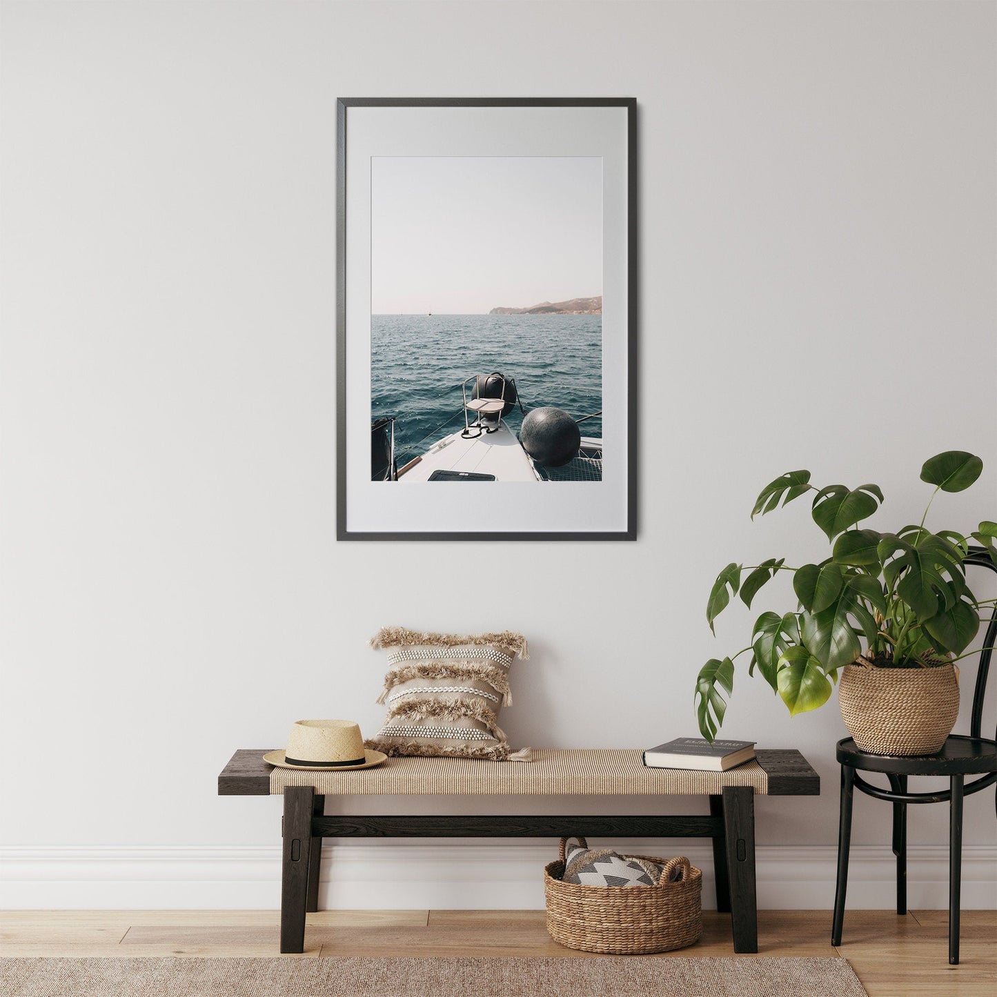 ‘Mediterranean Bliss’ framed picture of a boat sailing on the ocean hangs above a bench adorned with pillows, hat, and plant.
