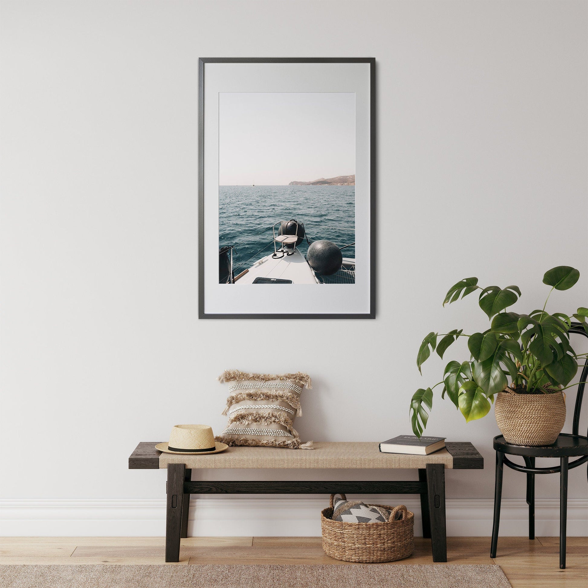 ‘Mediterranean Bliss’ framed picture of a boat sailing on the ocean hangs above a bench adorned with pillows, hat, and plant.