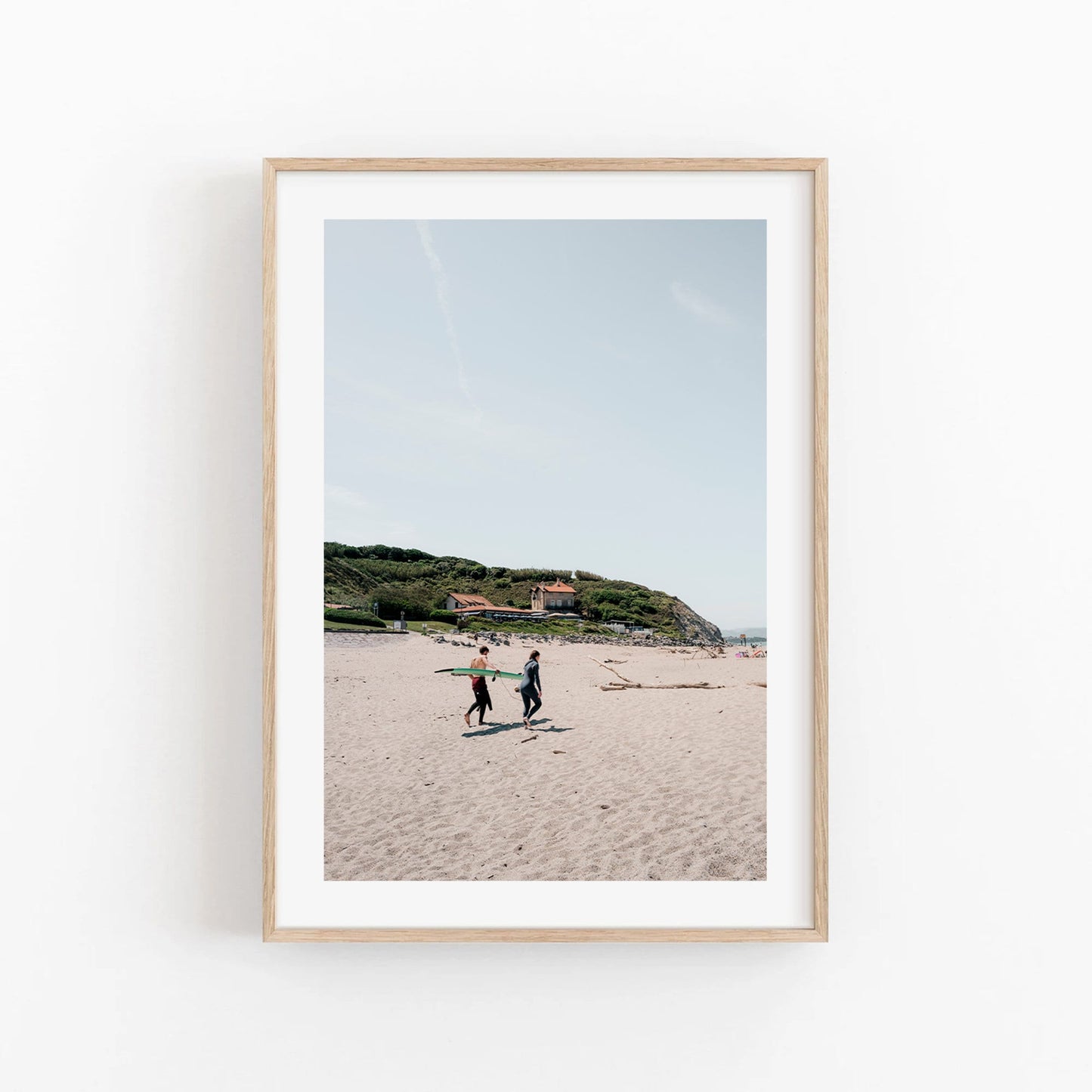Milady Surfers: A framed photo of two people walking on a sandy beach with hills and buildings in the background.