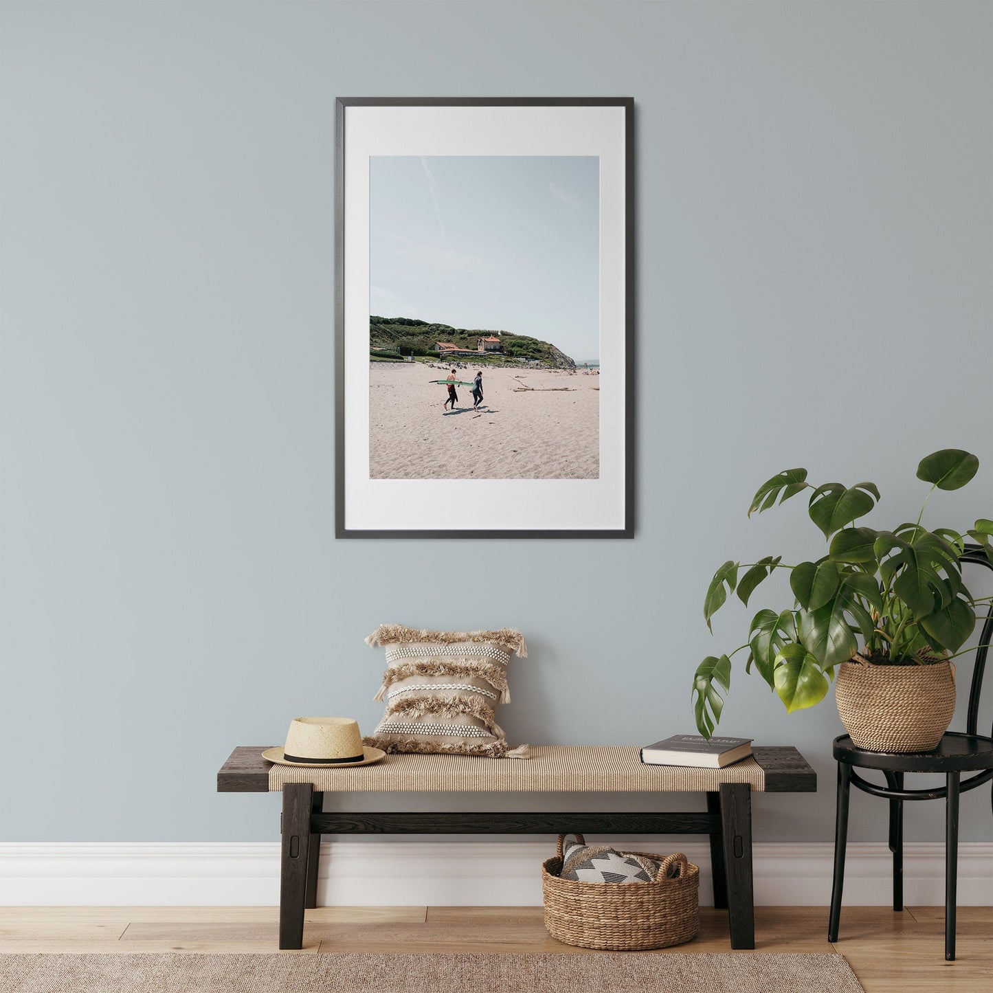 Milady Surfers beach photo on a light blue wall, above a bench with a pillow and hat, next to a potted plant.