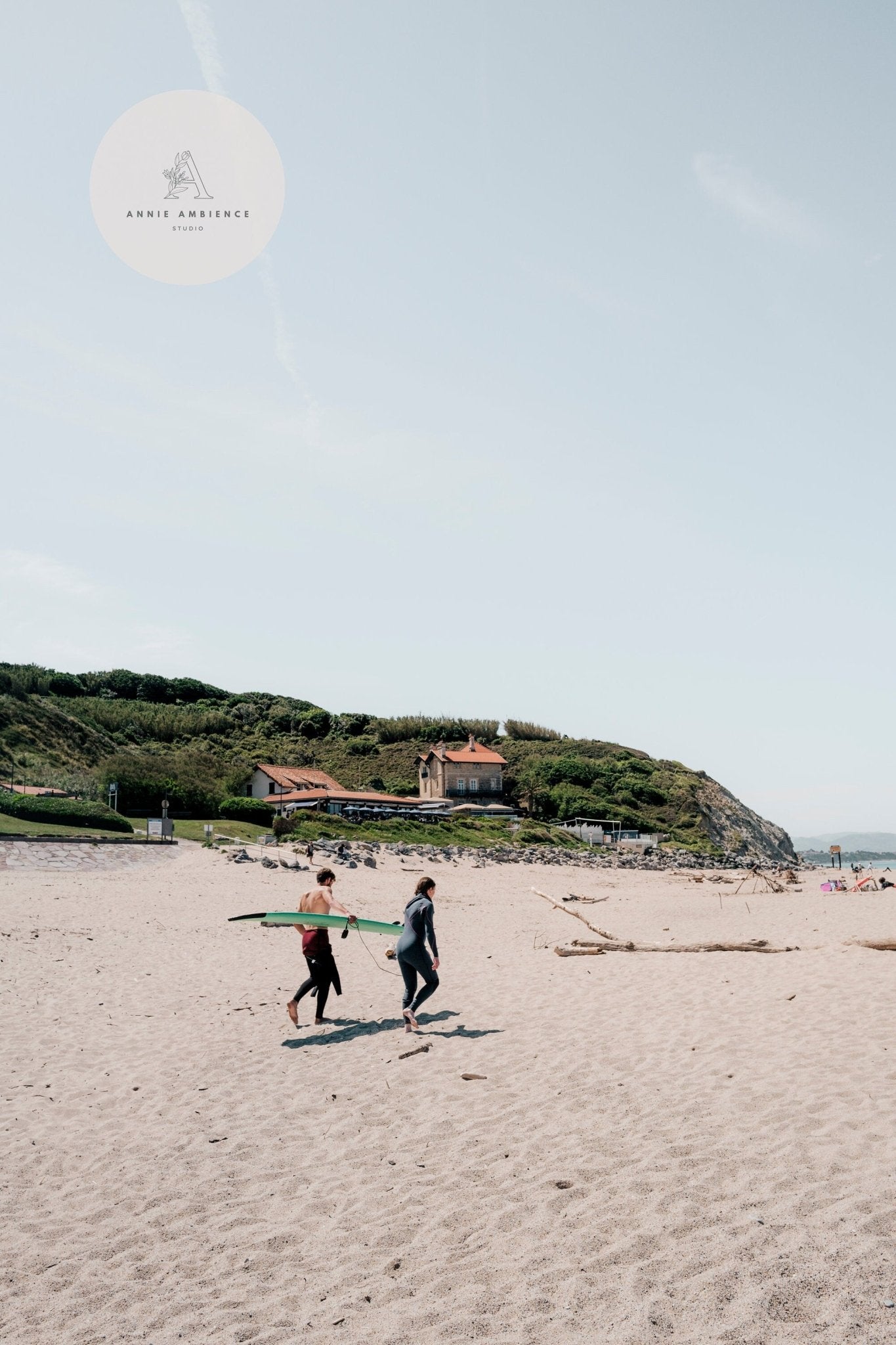 Two surfers walk on a sandy beach with surfboards, green cliffs, and houses behind them. #MiladySurfers.