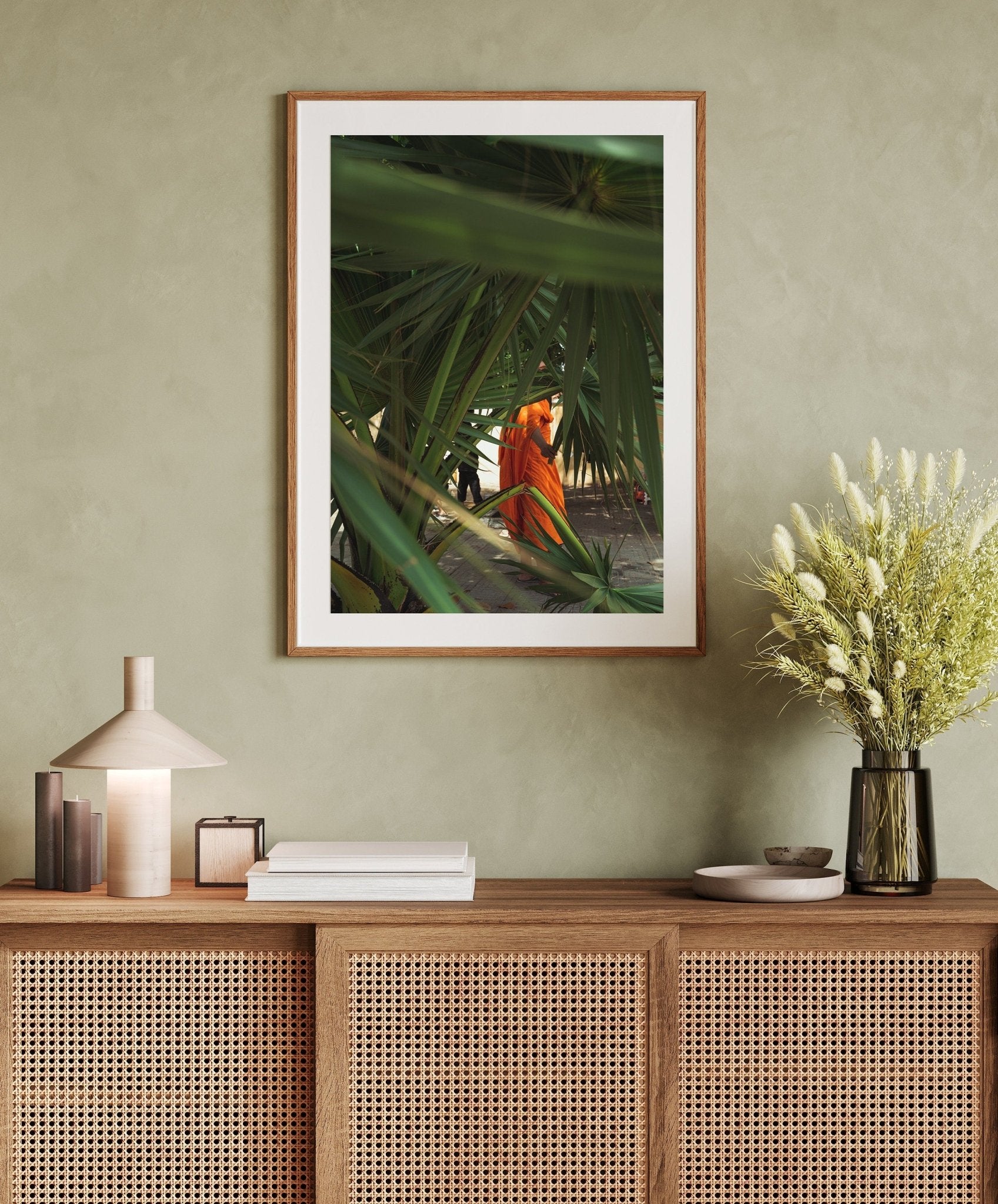 Monk framed art of person in orange amid greenery above a wicker cabinet with decor and flower vase.