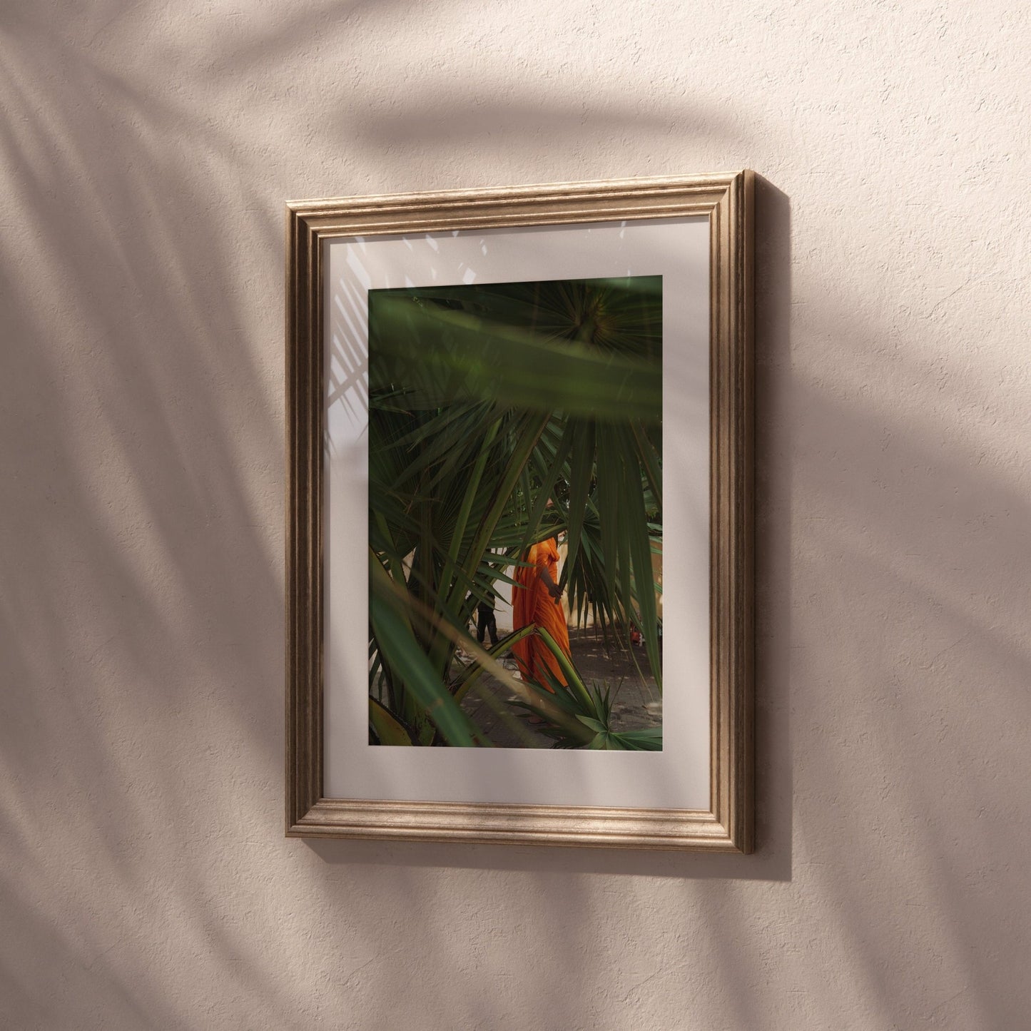 Framed Monk photo: palm leaves and a figure in orange on a textured beige wall.