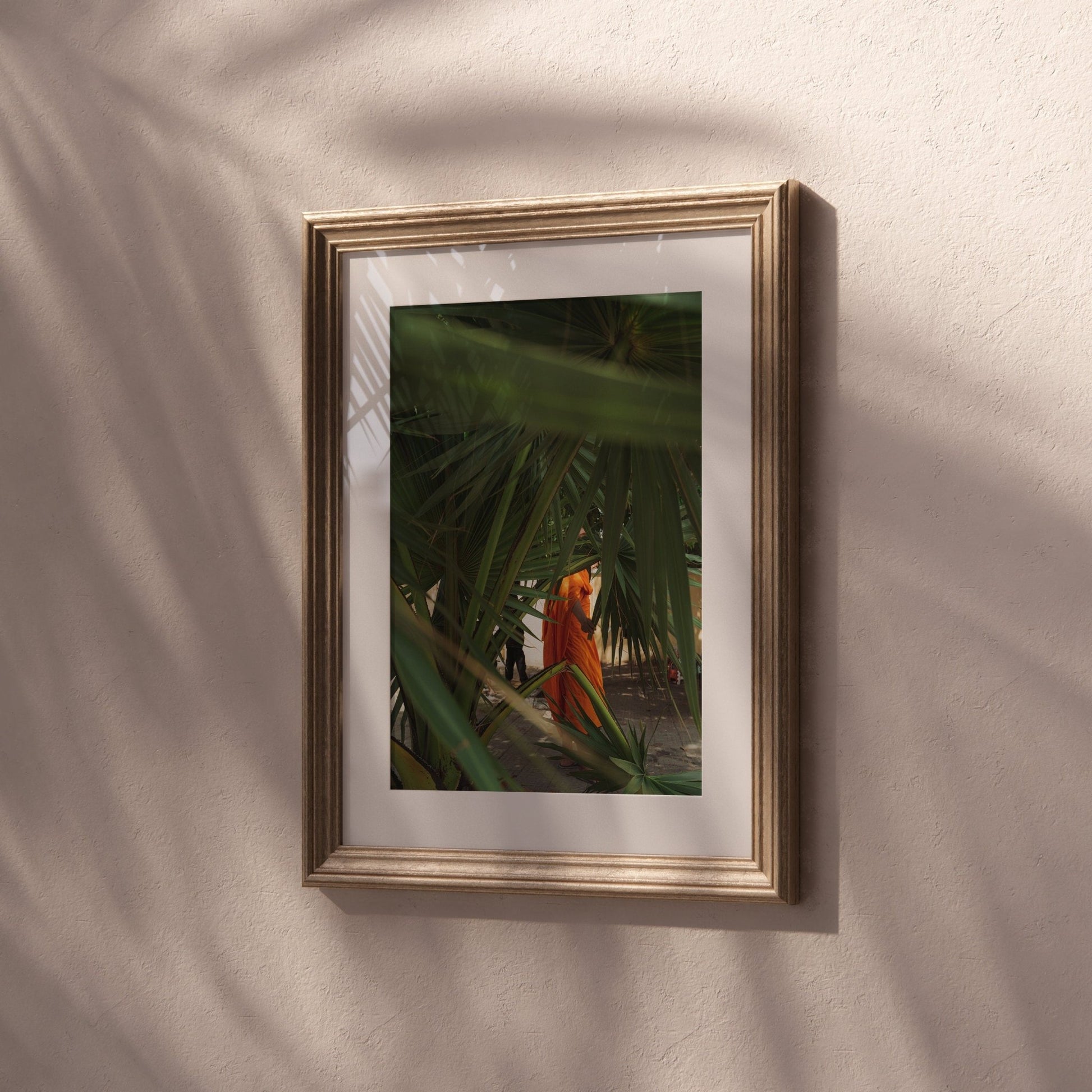 Framed Monk photo: palm leaves and a figure in orange on a textured beige wall.