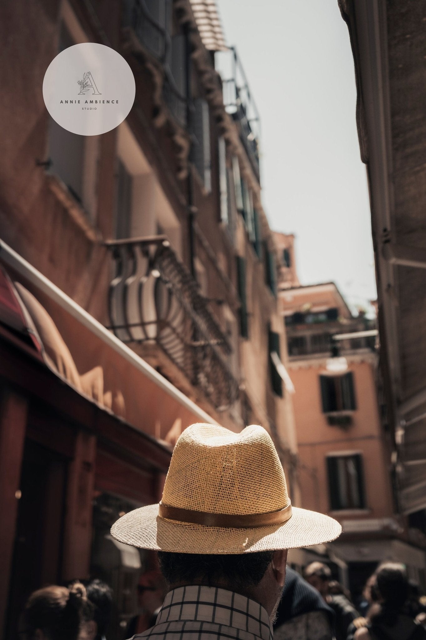 Mystery Man in a straw hat strolls through a sunlit, narrow street lined with tall buildings.
