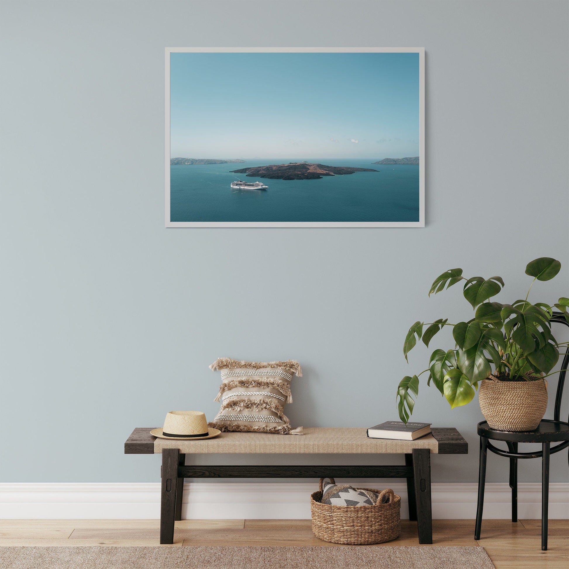 A room with a bench, hat, and plant featuring the Nea Kameni framed photo on a boat and island against a blue wall.