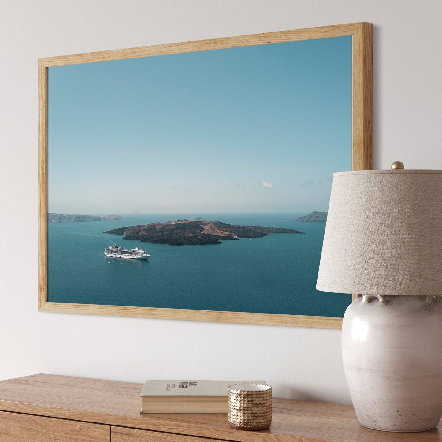 Nea Kameni framed seascape above a wooden table with a ceramic lamp and closed book.