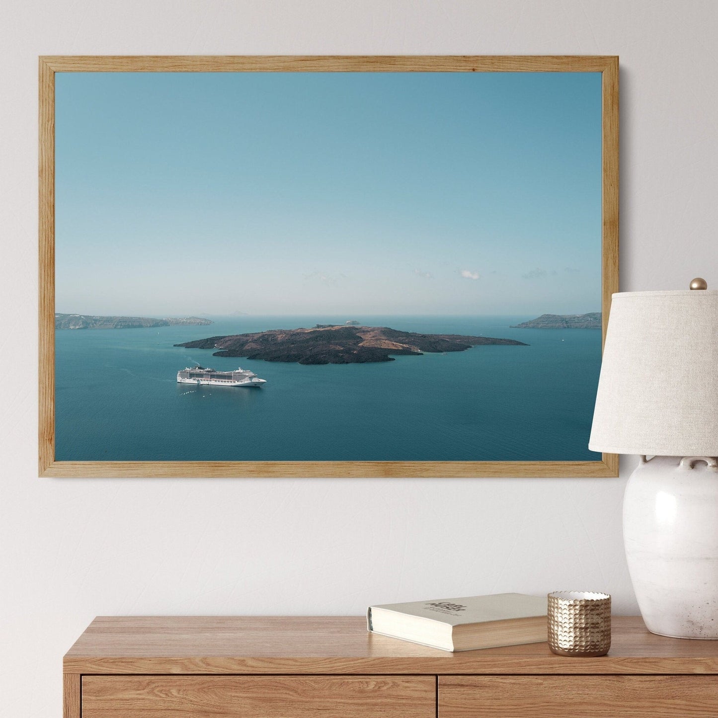 Framed image Nea Kameni of a cruise ship by an island, above a wooden cabinet with a lamp and book.