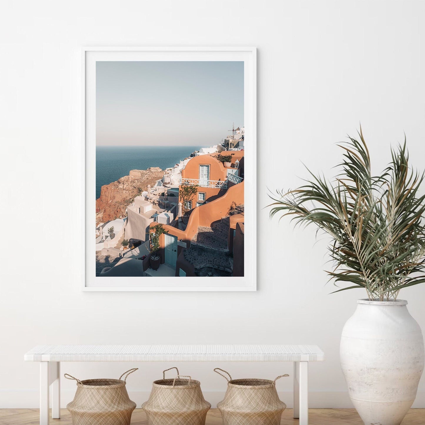 Oia framed photo of a coastal town on a white wall with a bench, baskets, and a potted plant.