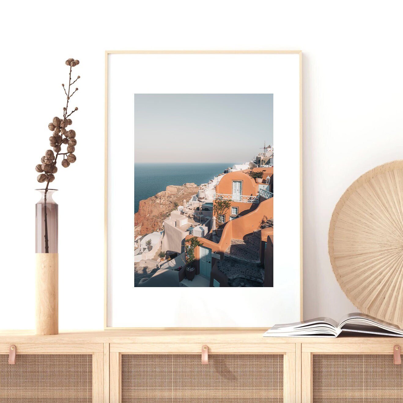 Oia scene photo in living room with vase, fan, and open book on wooden shelf.