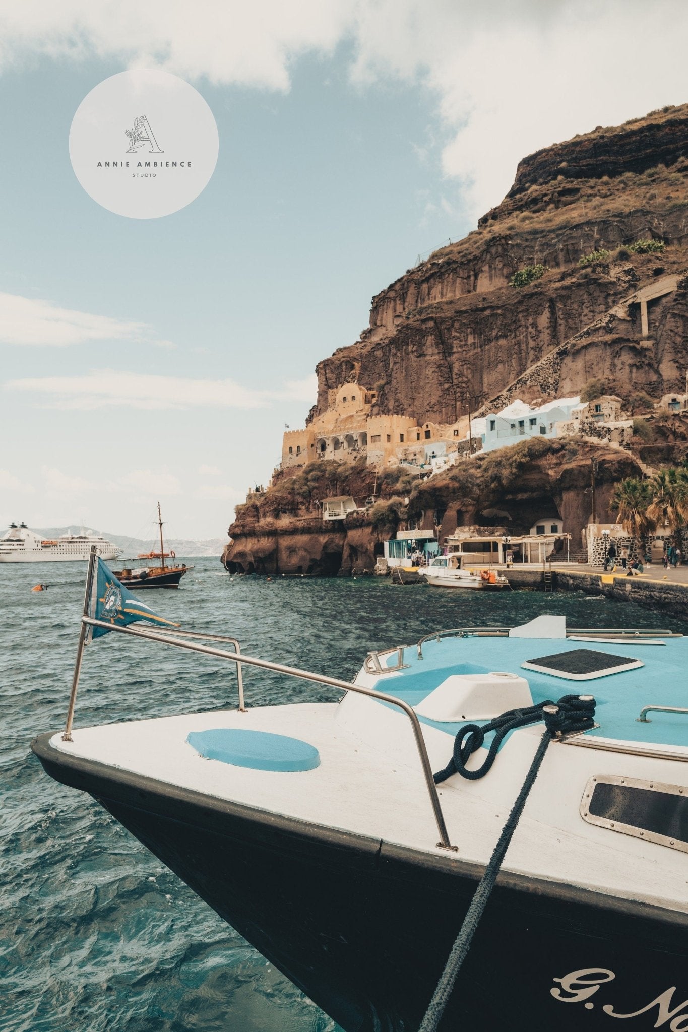 A white boat is docked in the foreground at the Old Port Fira, with rocky cliffs and buildings under a partly cloudy sky.
