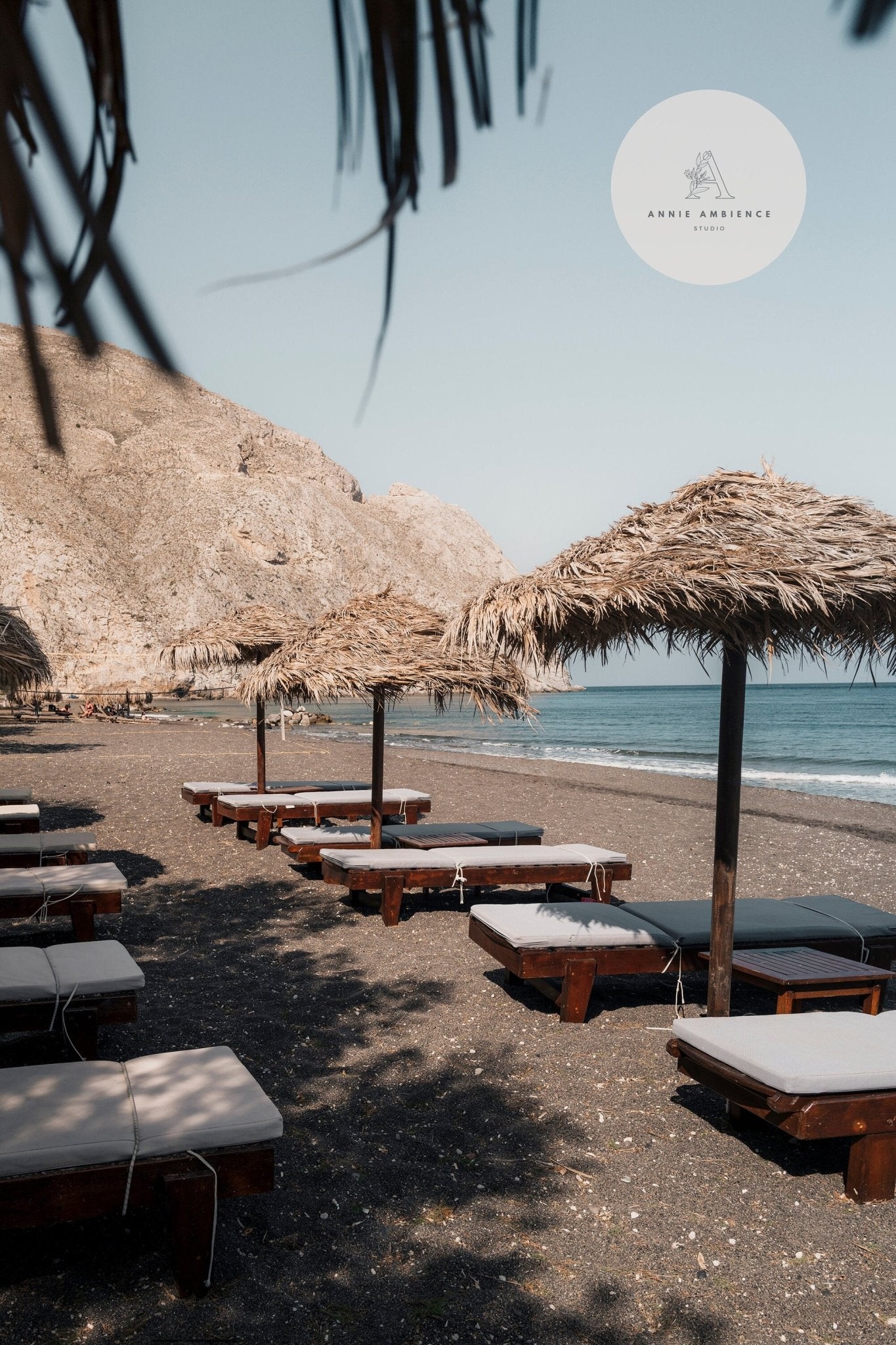 The Perissa - Set of 3 features a sandy beach with wooden sunbeds, straw umbrellas, rocky hills, and a calm sea.