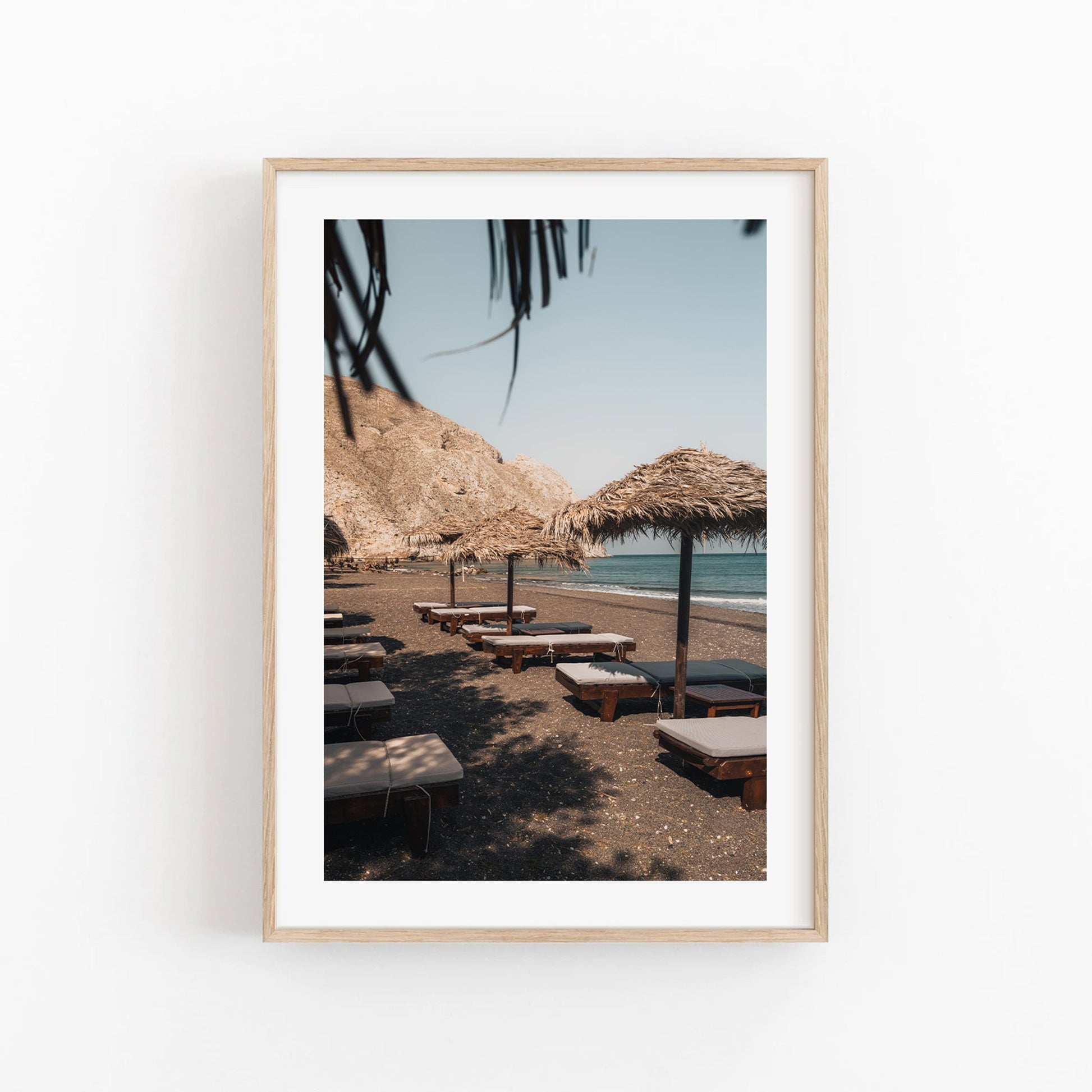 Framed Perissa Umbrellas photo: beach with lounge chairs, straw umbrellas, rocky hills, and a clear sky.