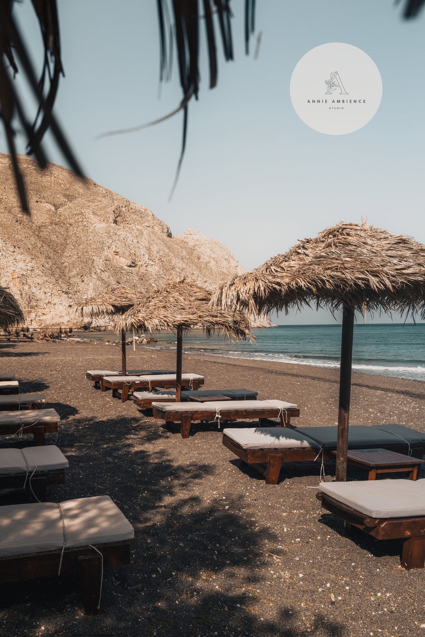 Perissa Umbrellas: sun loungers on a rocky beach, distant cliffs, and calm sea beneath a clear sky.