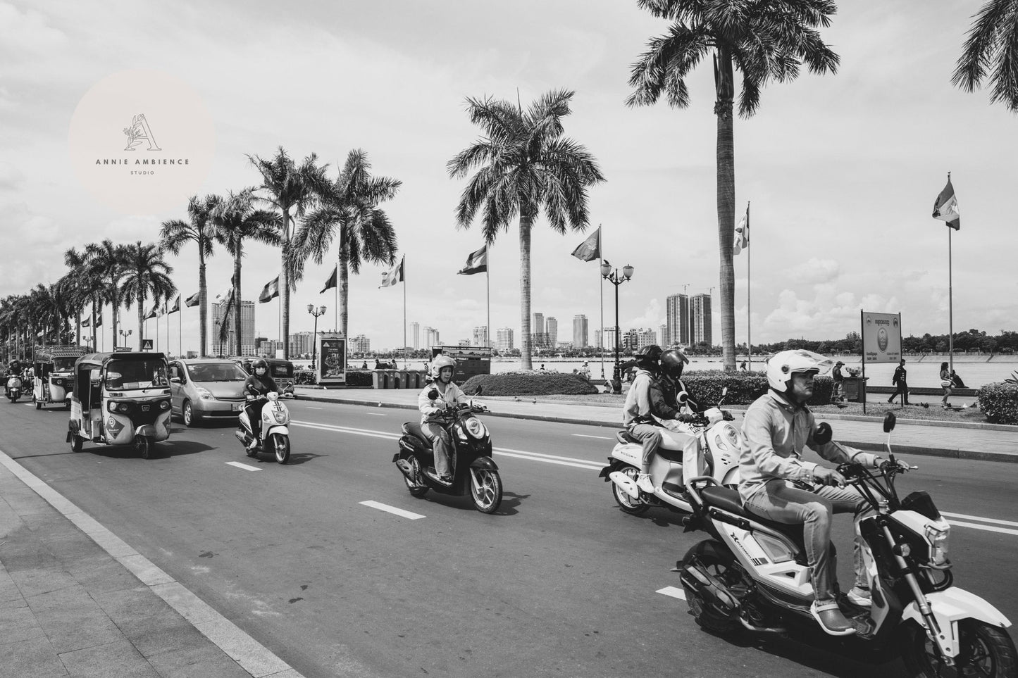 Riding scooters and vehicles on a palm-lined street near a city skyline, captured in Phnom Penh Black and White.