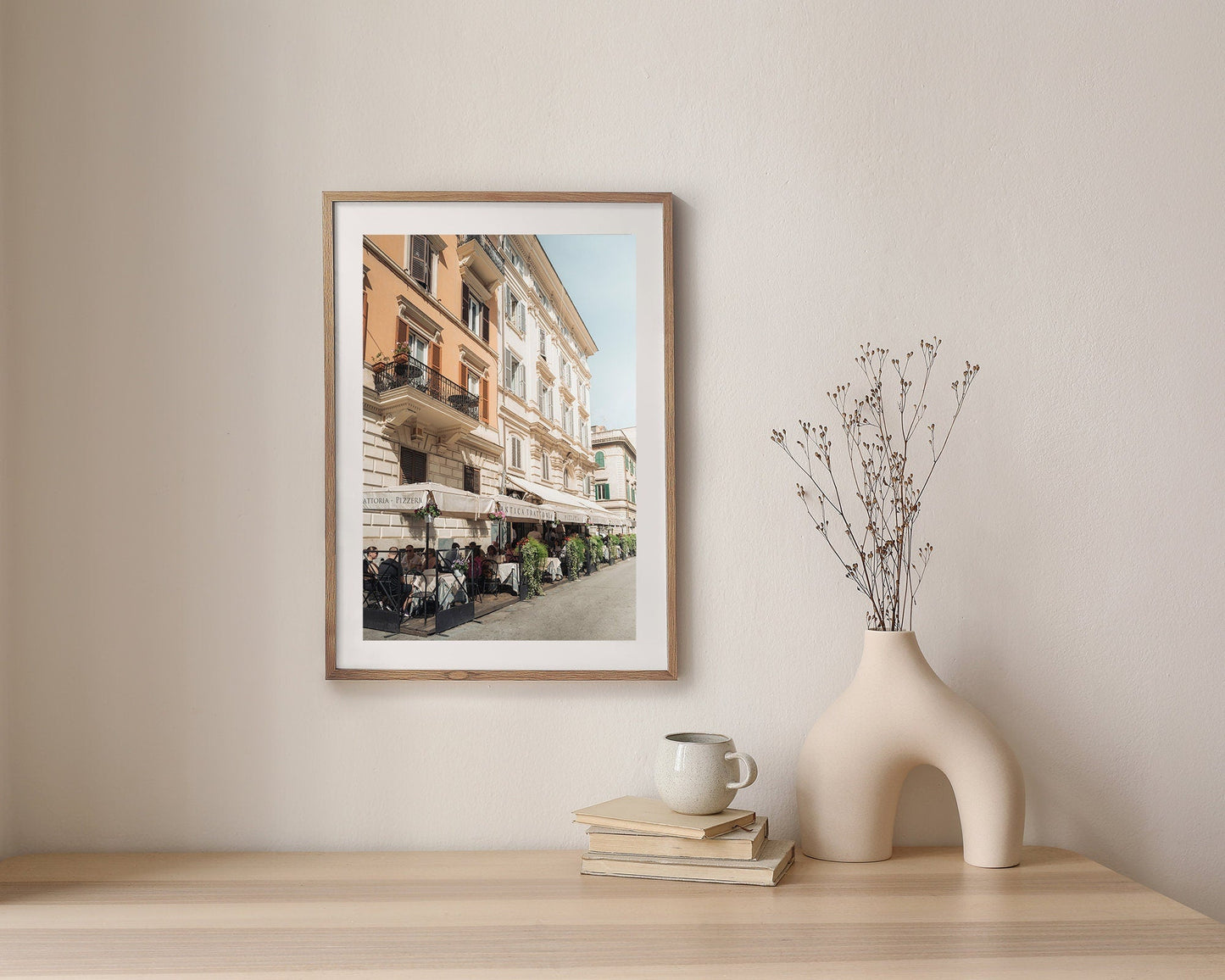 Framed Pizzeria photo on a wall beside a sculptural vase, dried branches, cup, and books on a wooden table.