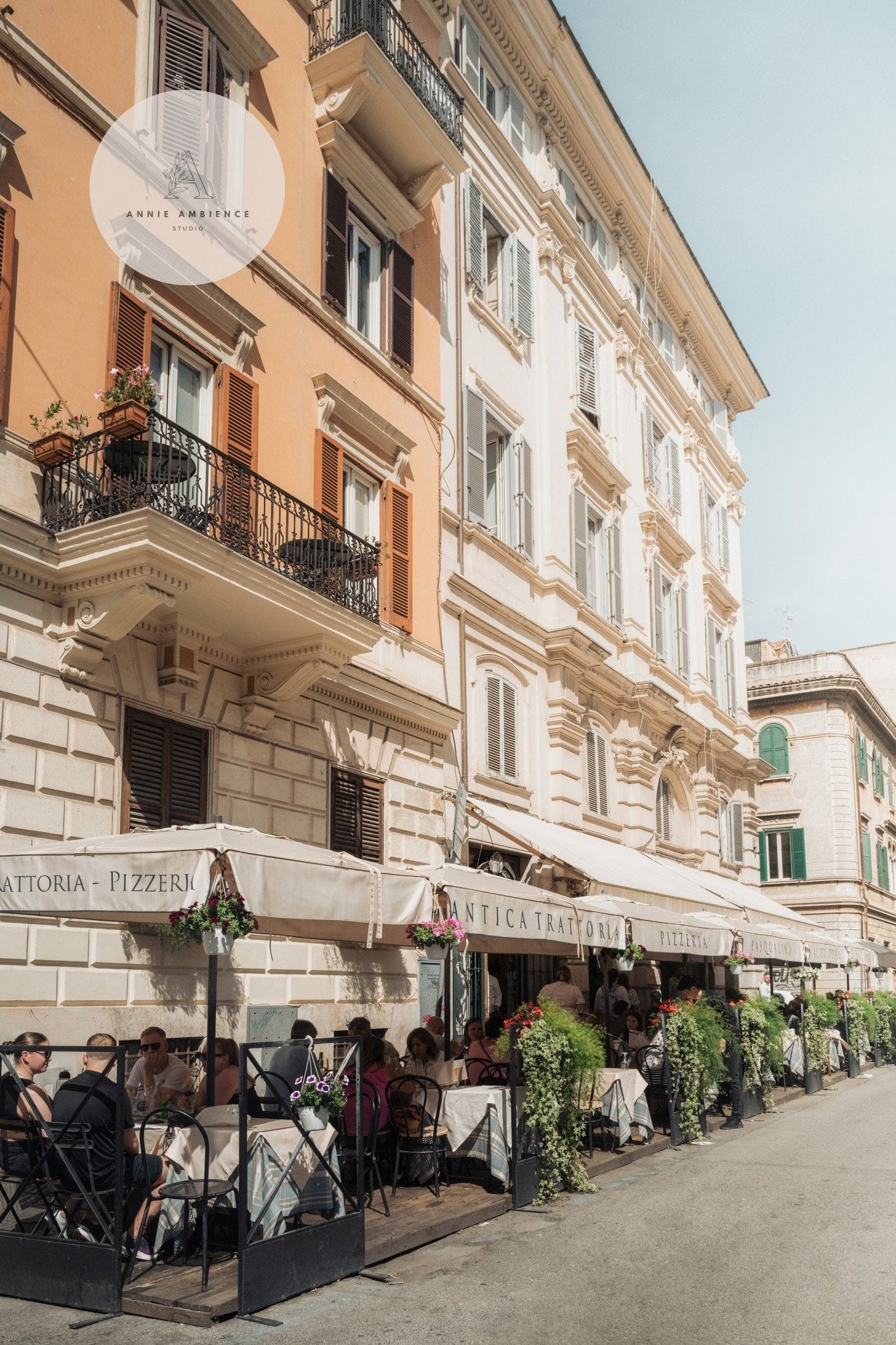 European street with people dining under canopies at a Pizzeria, flowers decorating the sidewalk.