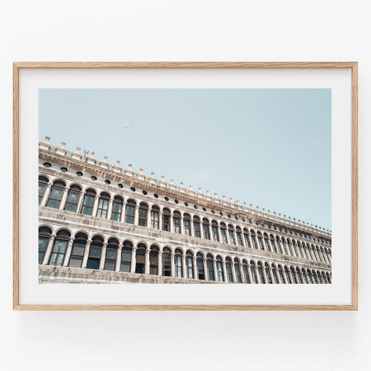Framed photo of the Procuratie Vecchie, an ornate historic building with columns and arches against a clear blue sky.