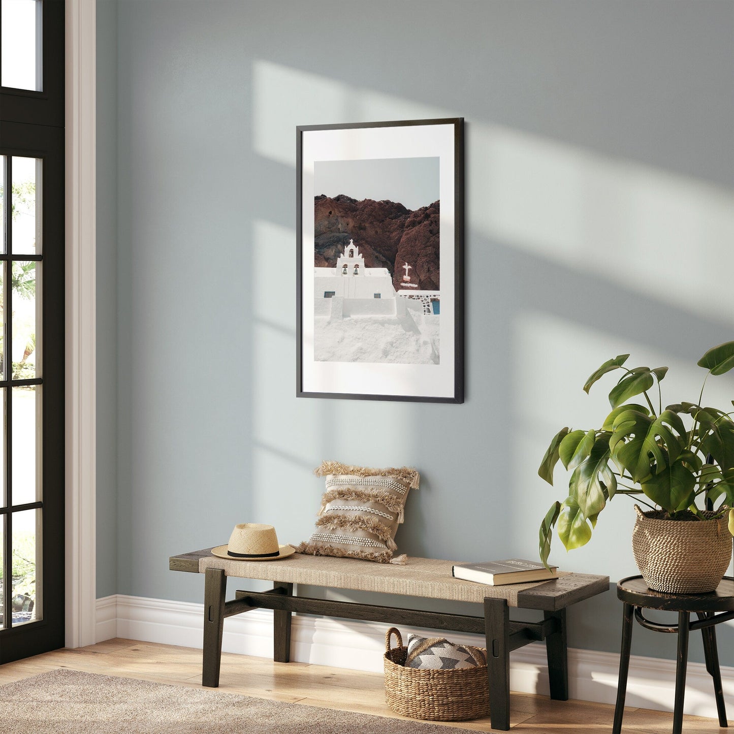 A sunlit room with a bench, pillow, and hat; Red Beach Church framed on the wall and a plant in the corner.