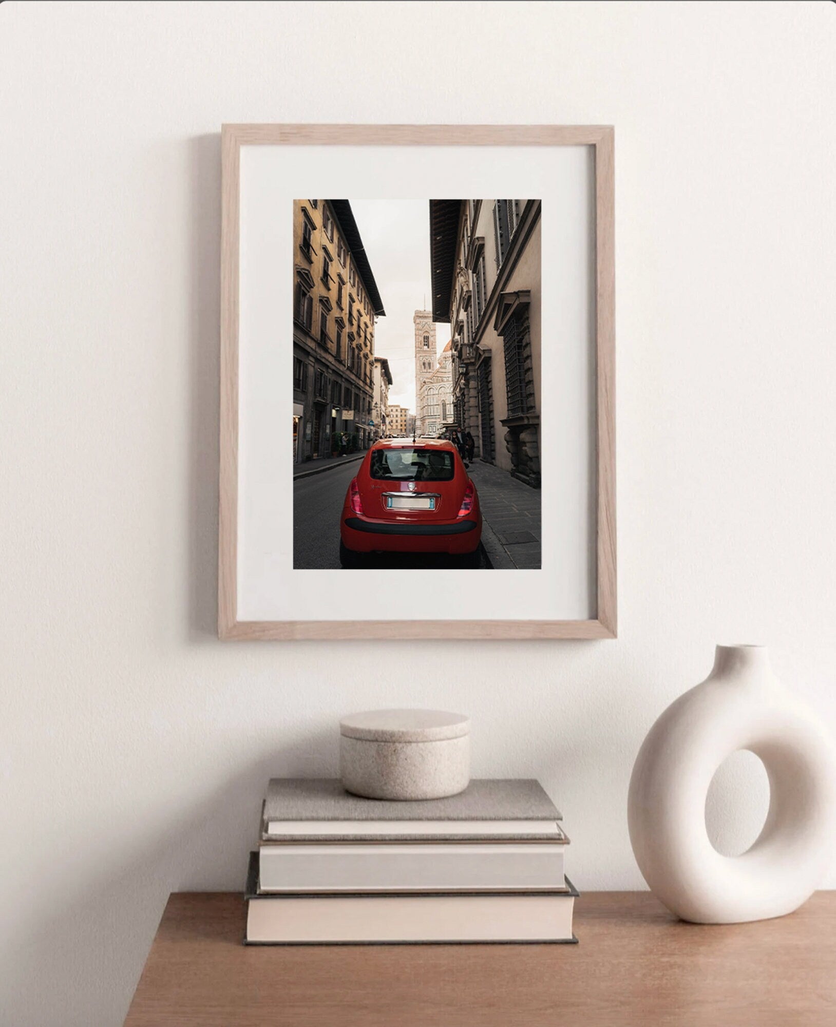 Framed Red Car photo on city street, above books and vase on a wooden table.