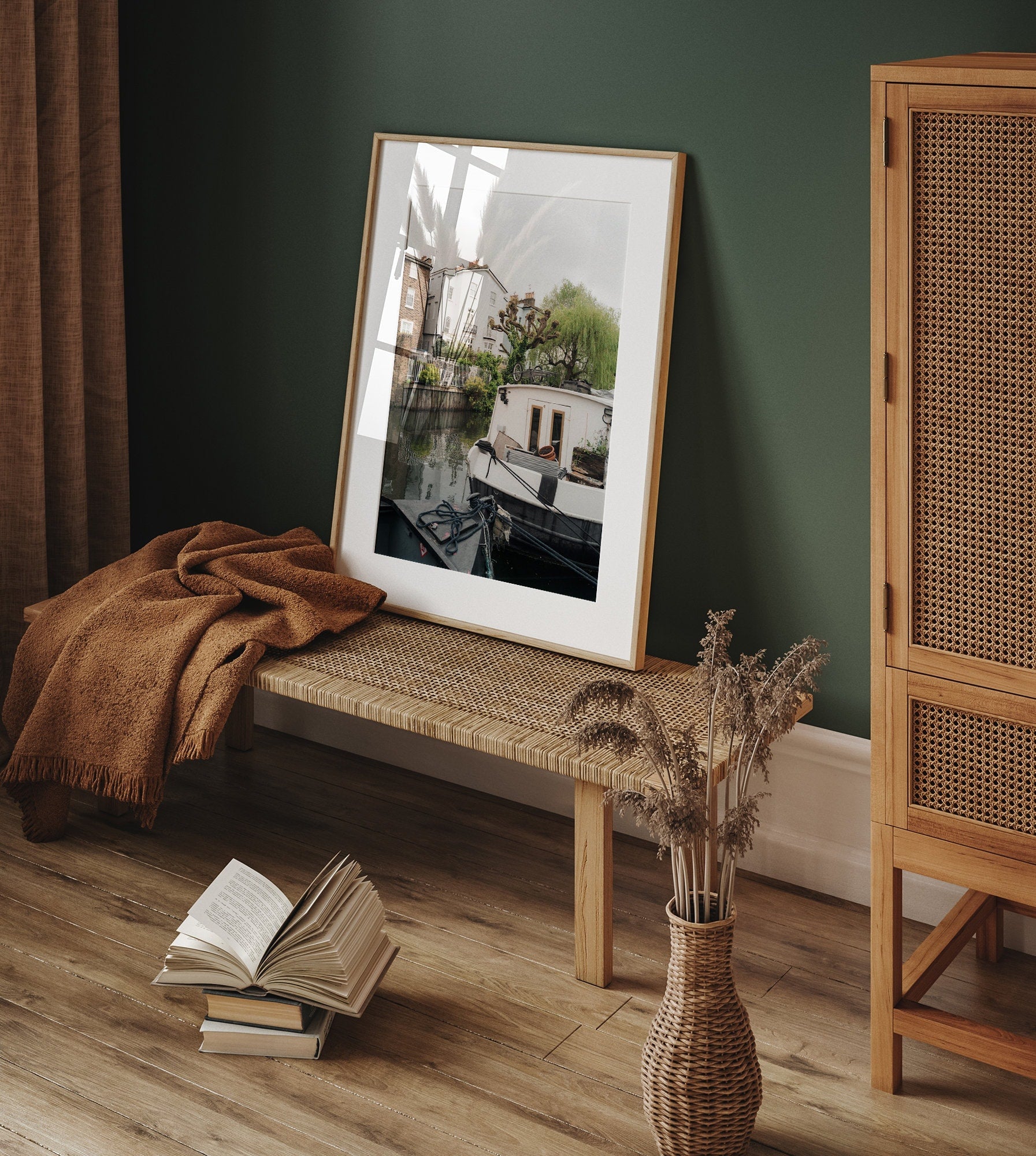 Framed Regents Canal II photo on woven bench; open book, throw blanket nearby, vase of dried flowers on wooden floor.