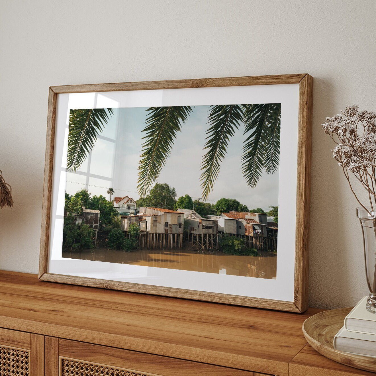 Framed River Homes photo with palm leaves on a wooden cabinet near dried flowers.