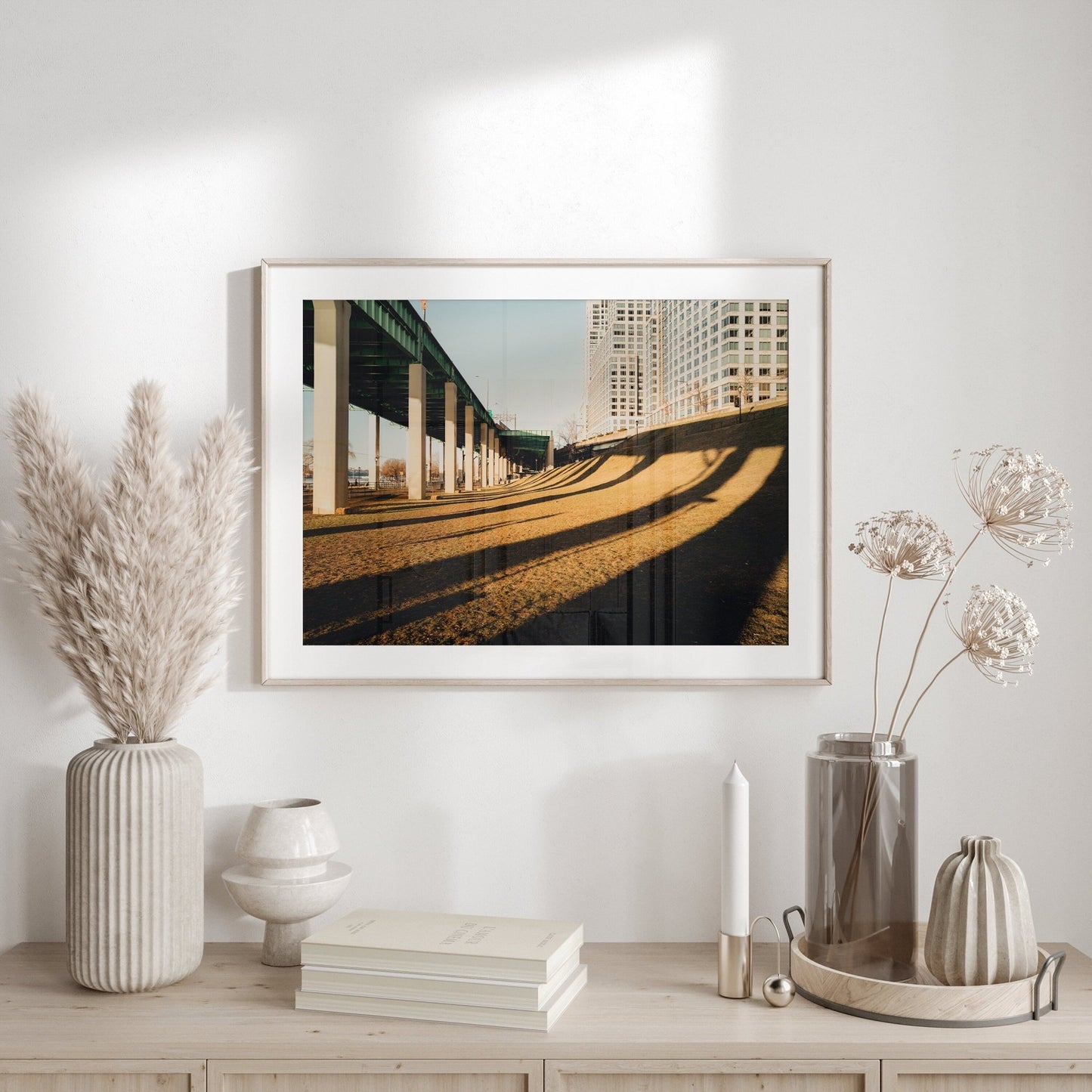 The Riverside framed picture of a bridge casts shadows, displayed on a shelf with pampas grass, vases, and a candle.