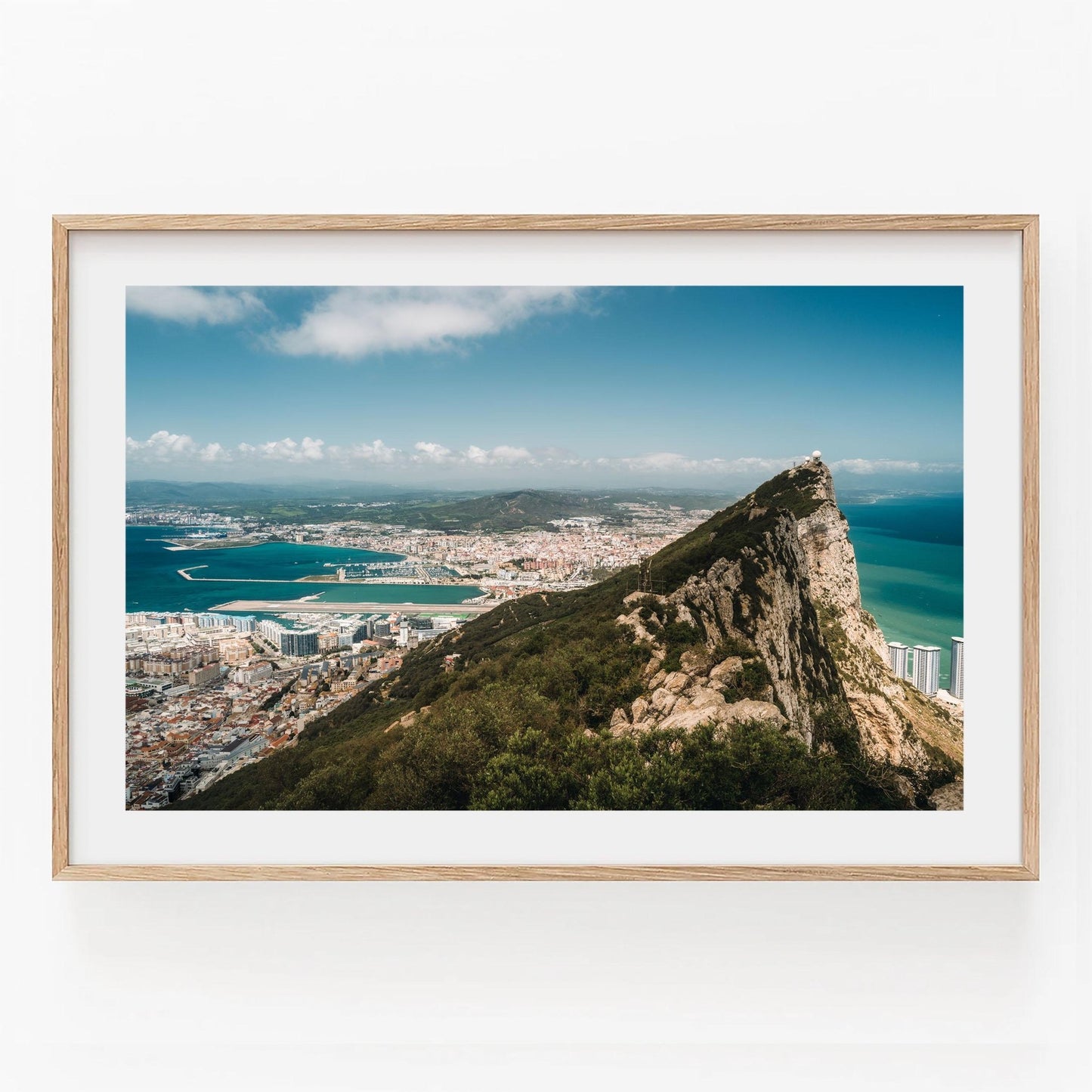 Framed Rock of Gibraltar photo: coastal city view with mountains, blue sea, clear sky, and rocky terrain foreground.