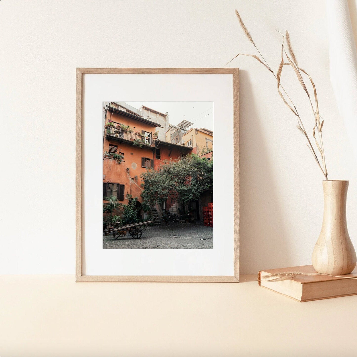 The Rome Courtyard framed image of a rustic courtyard with orange walls is displayed on a shelf with a vase and books.