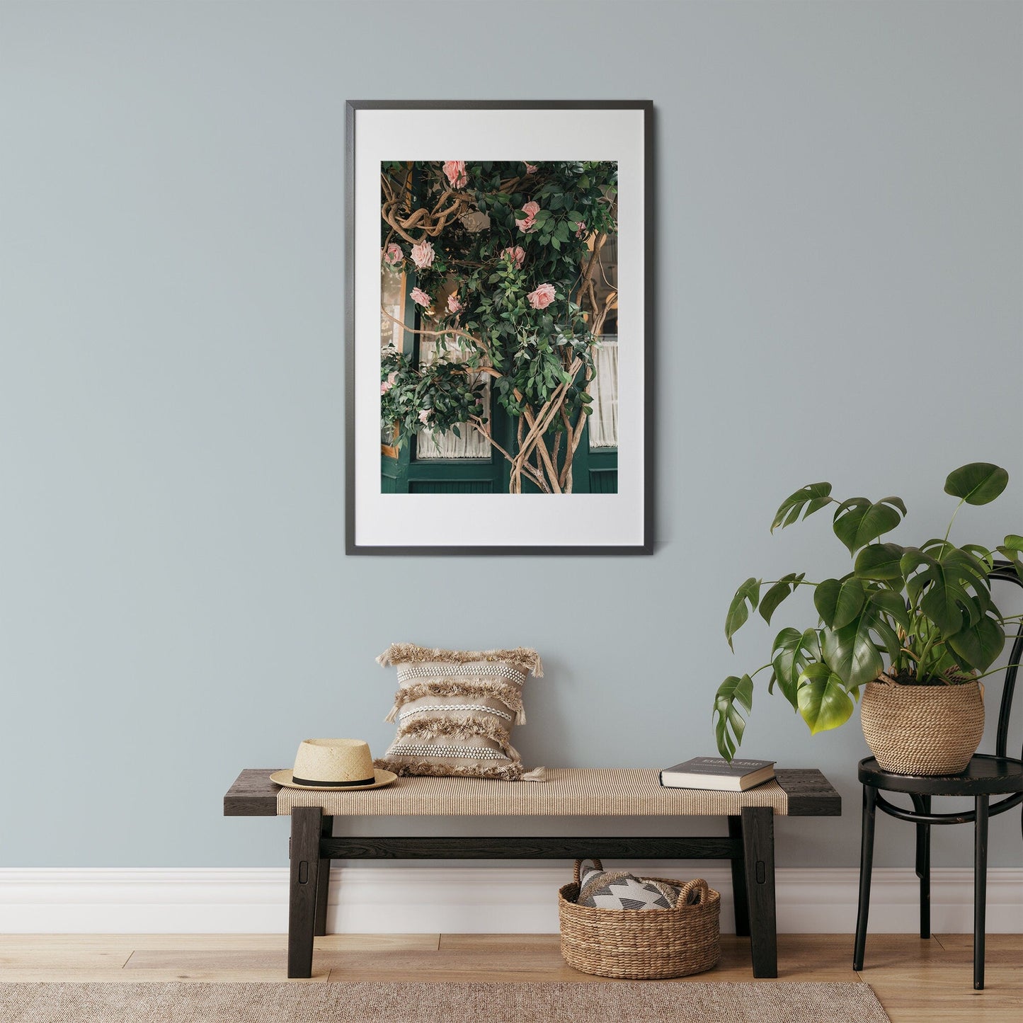 The Rose Vine framed artwork hangs on a light gray wall above a bench with a pillow, hat, and nearby potted plant.