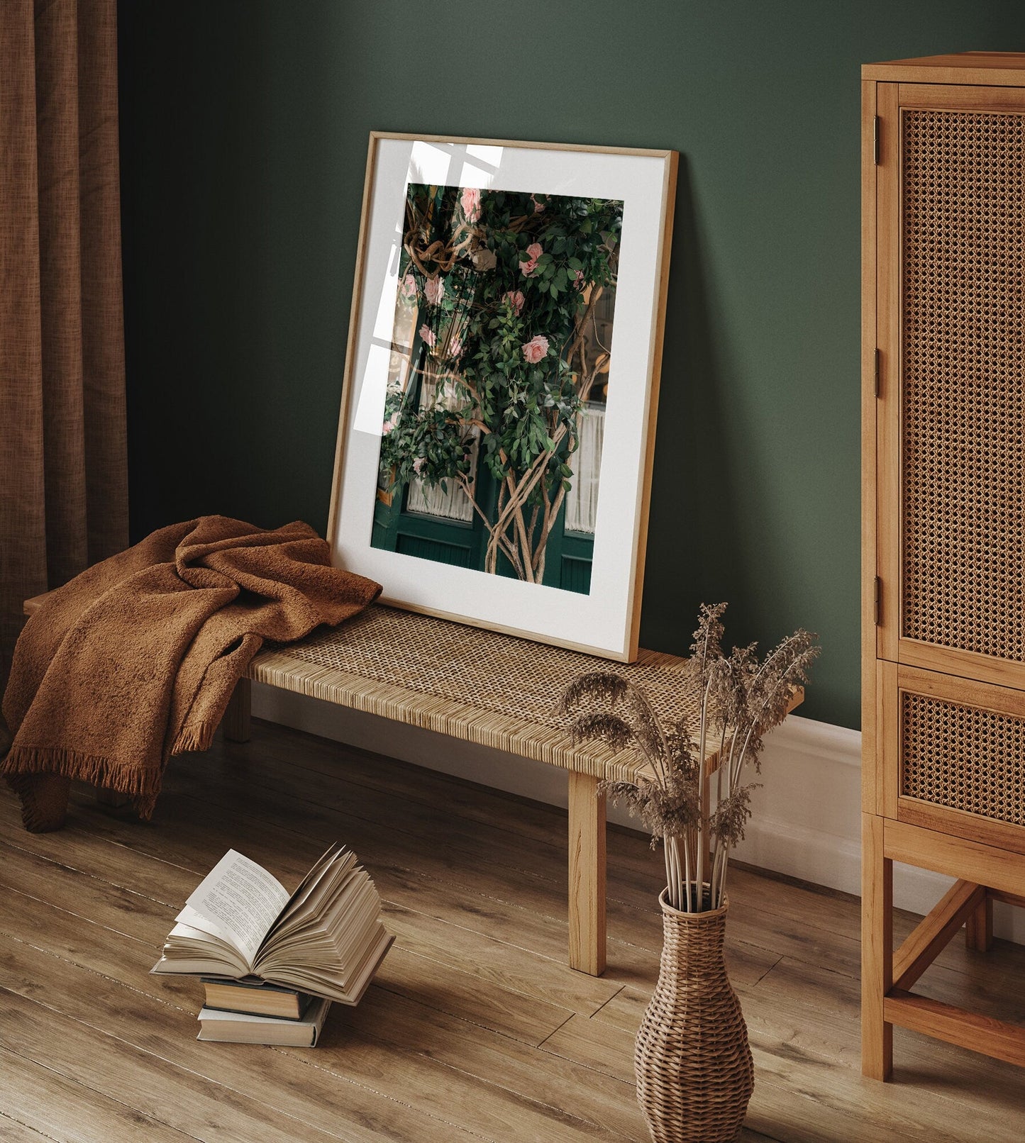 A cozy room with a wicker bench, Rose Vine artwork, open book, blanket, and dried plants in a vase.