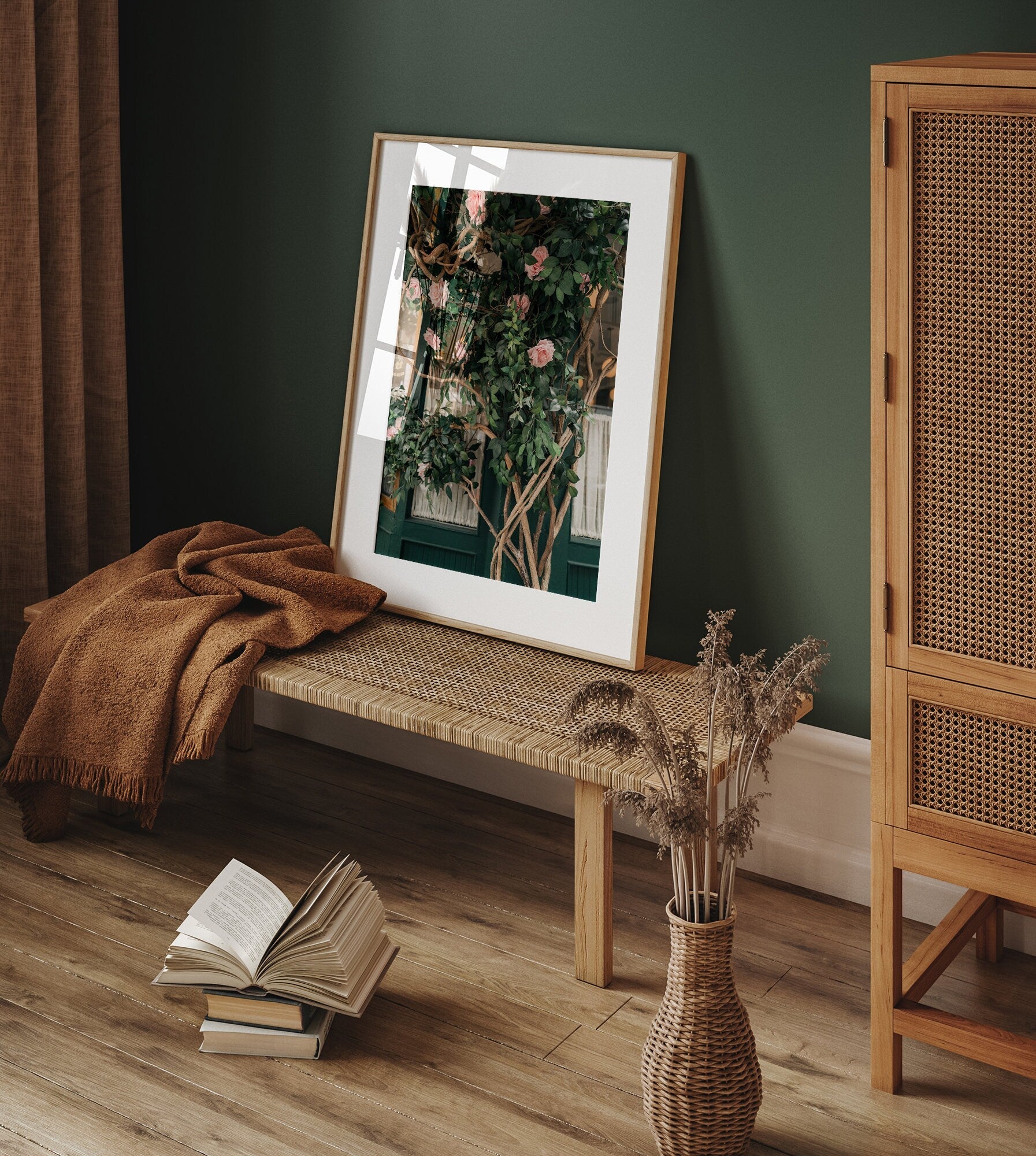 A cozy room with a wicker bench, Rose Vine artwork, open book, blanket, and dried plants in a vase.