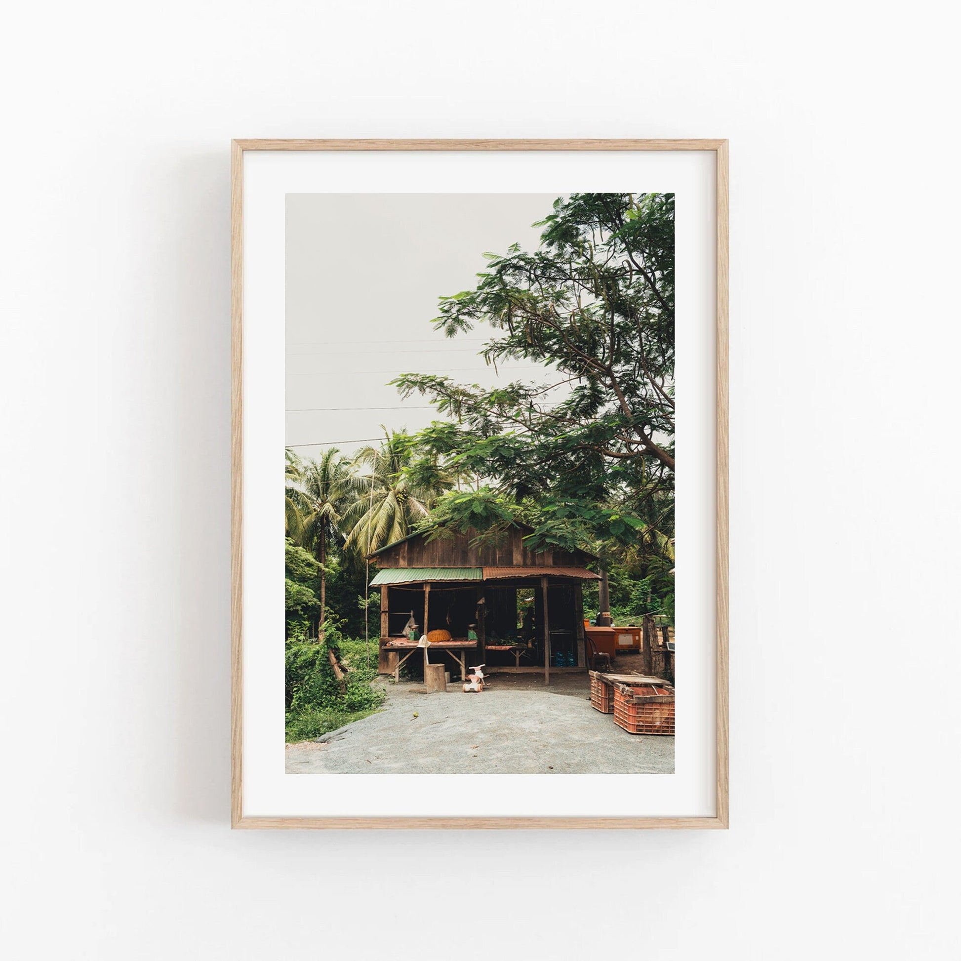Framed photo titled Rural Shop featuring a small wooden hut with surrounding trees and plants on a light background.