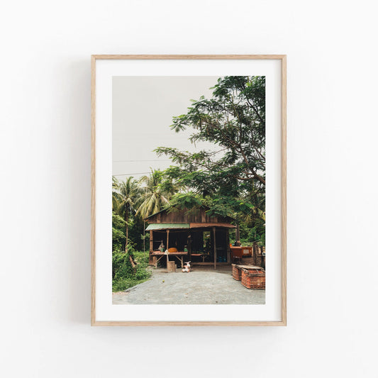 Framed photo titled Rural Shop featuring a small wooden hut with surrounding trees and plants on a light background.