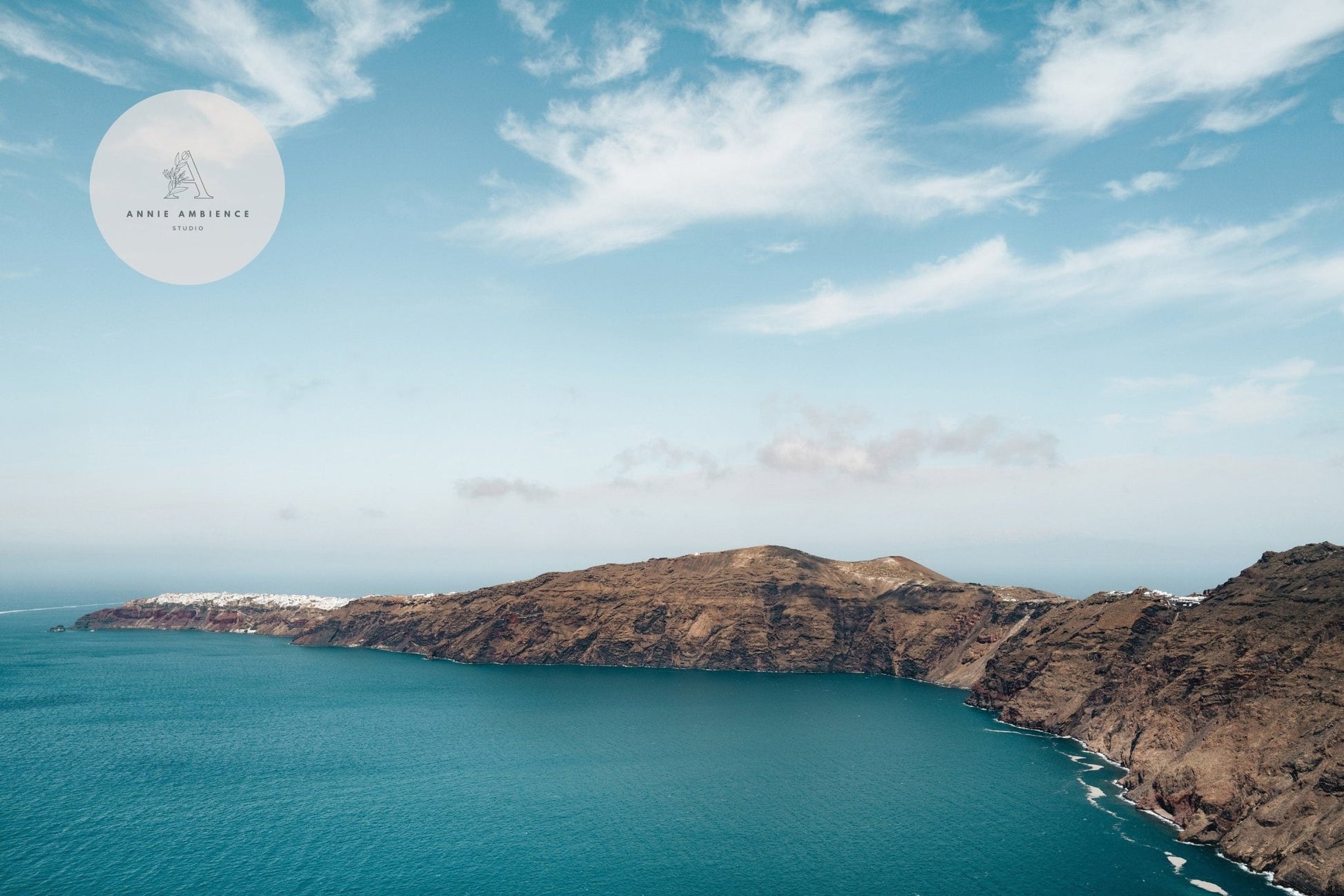 Aerial view of a rocky coast with a turquoise sea under clouds, Santorini Mountain logo in the top left corner.