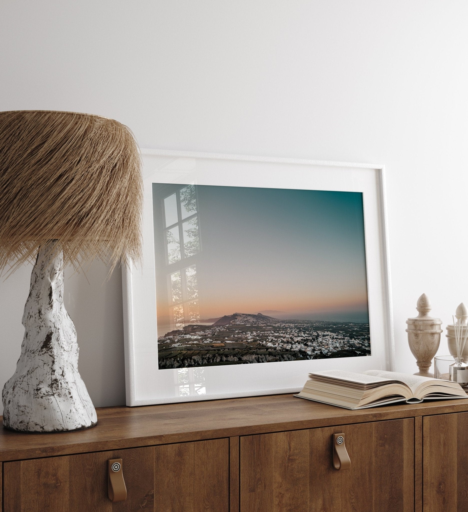 Santorinis Glow photo in a frame on a wooden cabinet with a lamp and an open book.