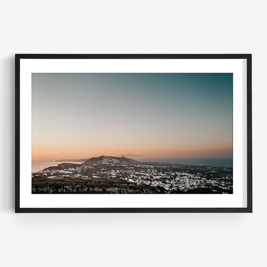 Santorinis Glow: A framed photo of a coastal town at sunset with white buildings and a hilly landscape.
