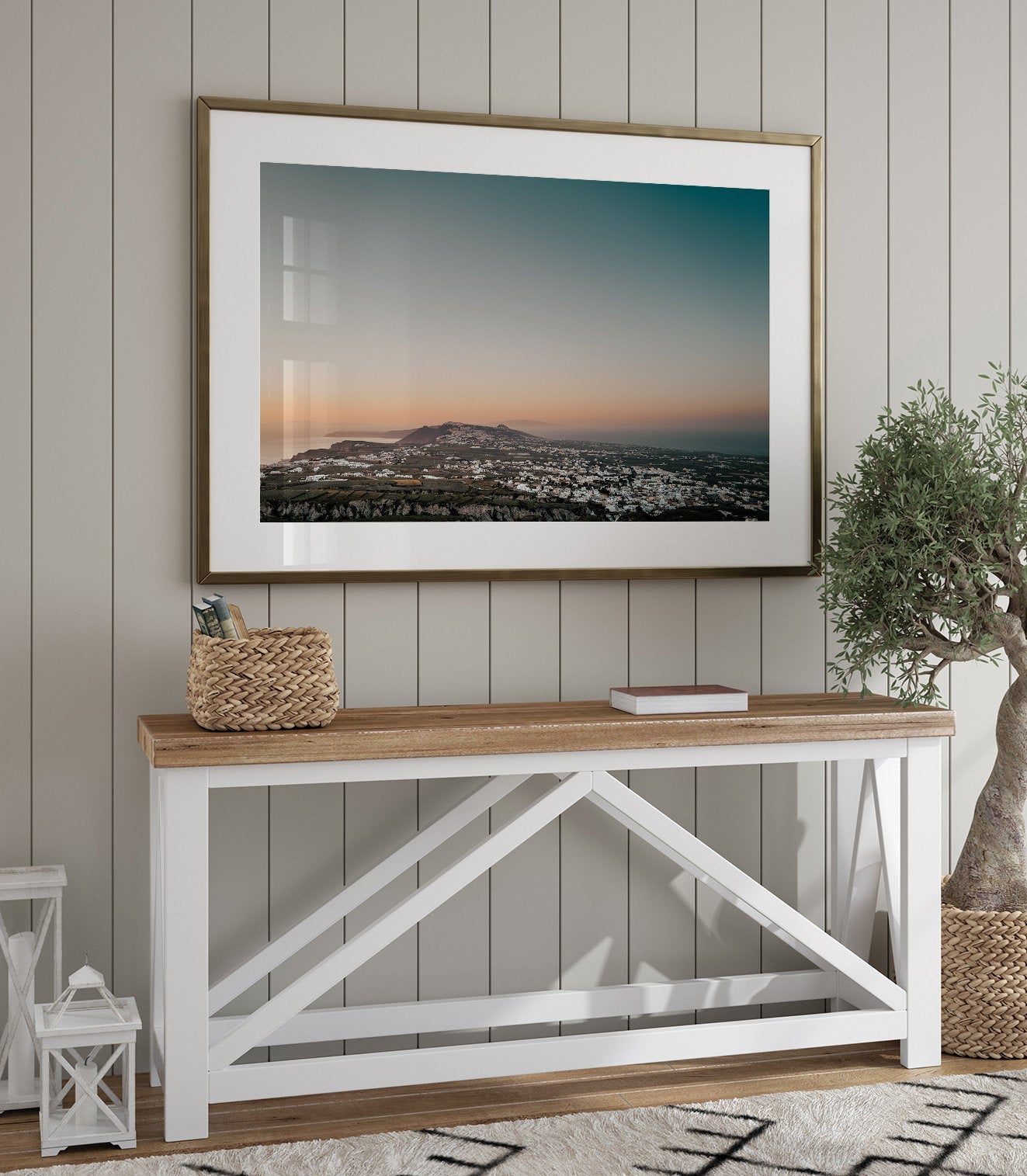 A modern living room with Santorinis Glow, a framed landscape photo, wooden console table, basket, book & potted plant.