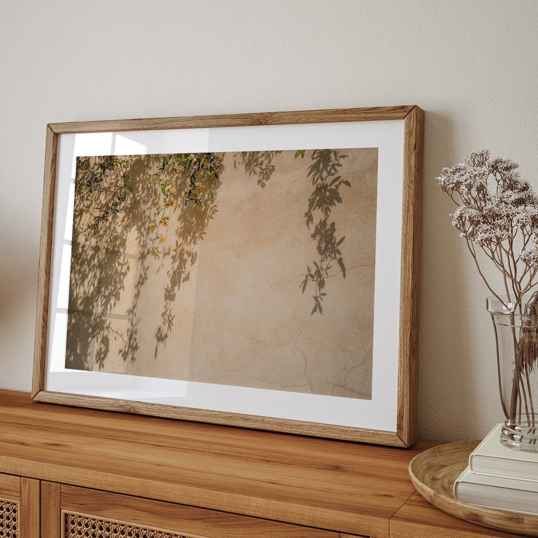 Framed Spring Breeze artwork on a wooden table, plant shadows on wall, beside a vase with dried flowers.
