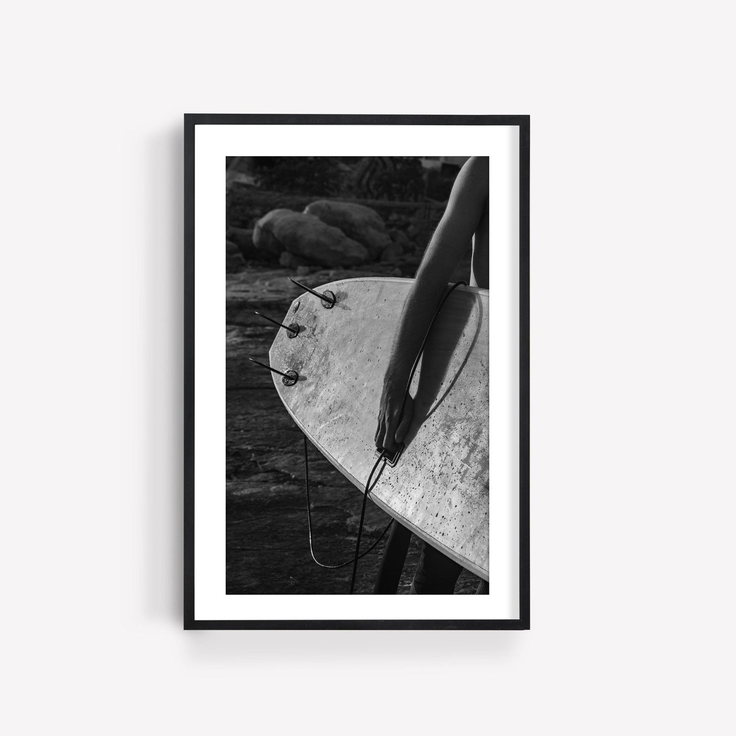 A person holds a surfboard in the black and white Surf Spirit photo with rocks behind them.
