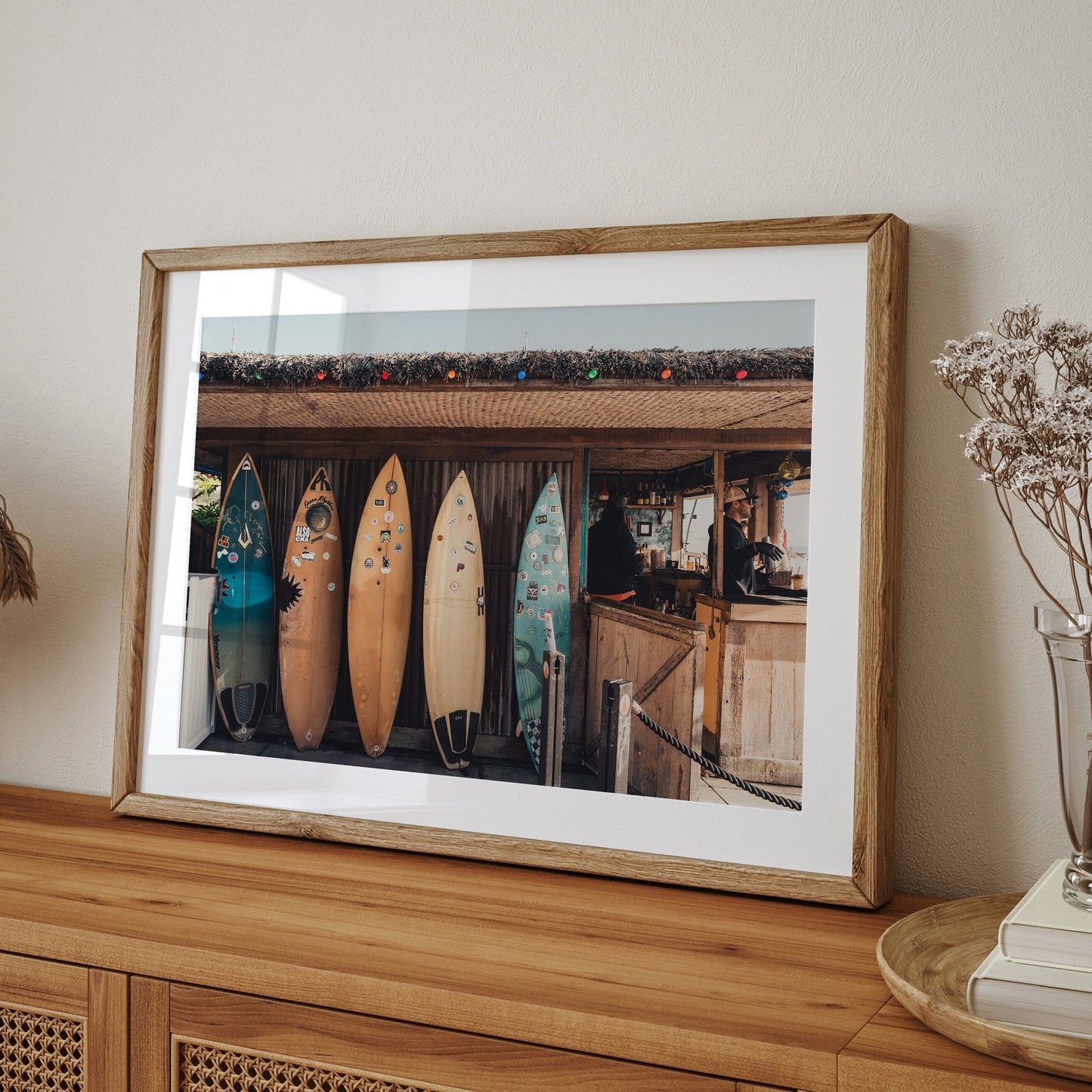 Framed Surfboard Cafe photo on a wooden sideboard with dried flowers, showcasing colorful surfboards by a rustic hut.