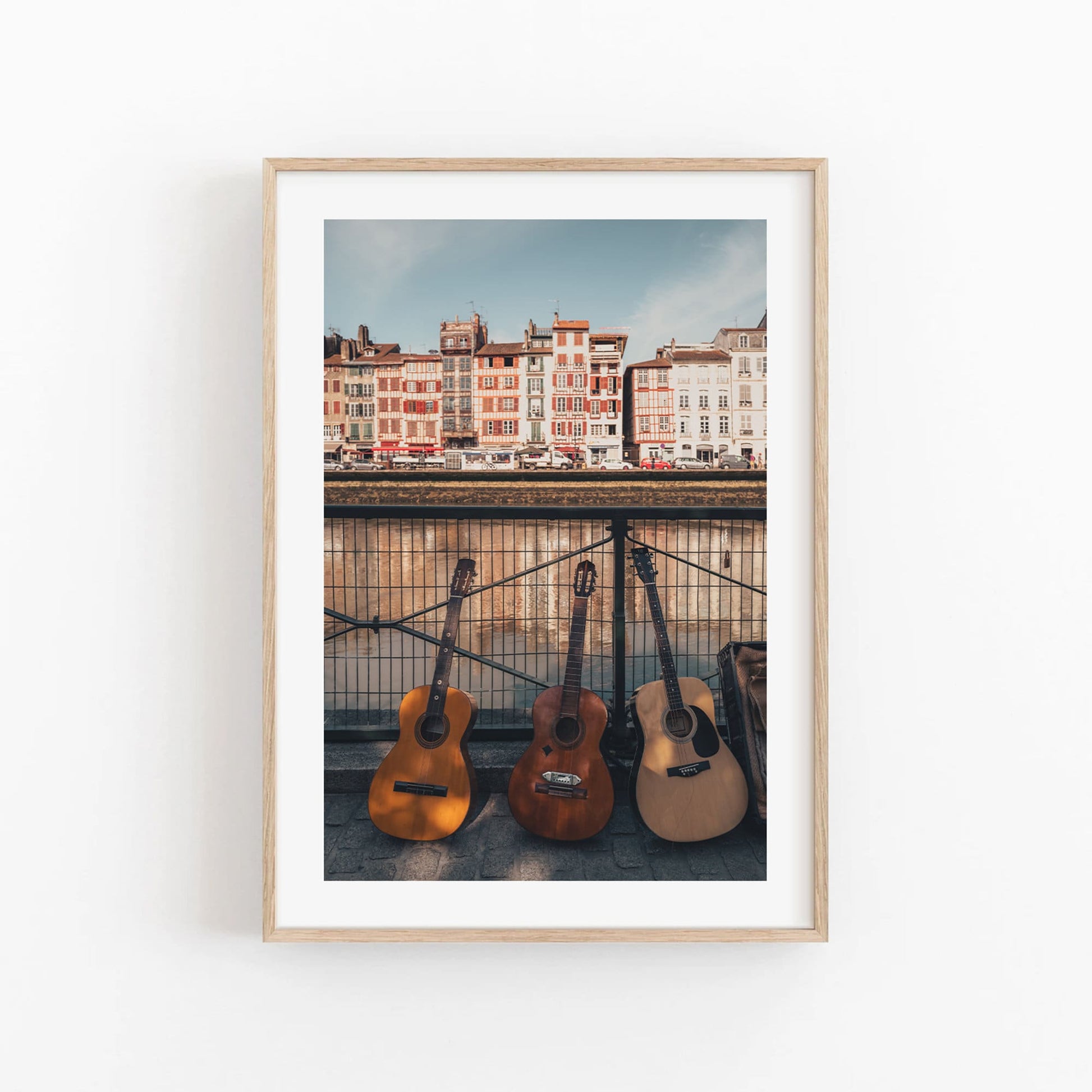 Framed photo of three guitars leaning against a waterfront railing with colorful buildings in the background.